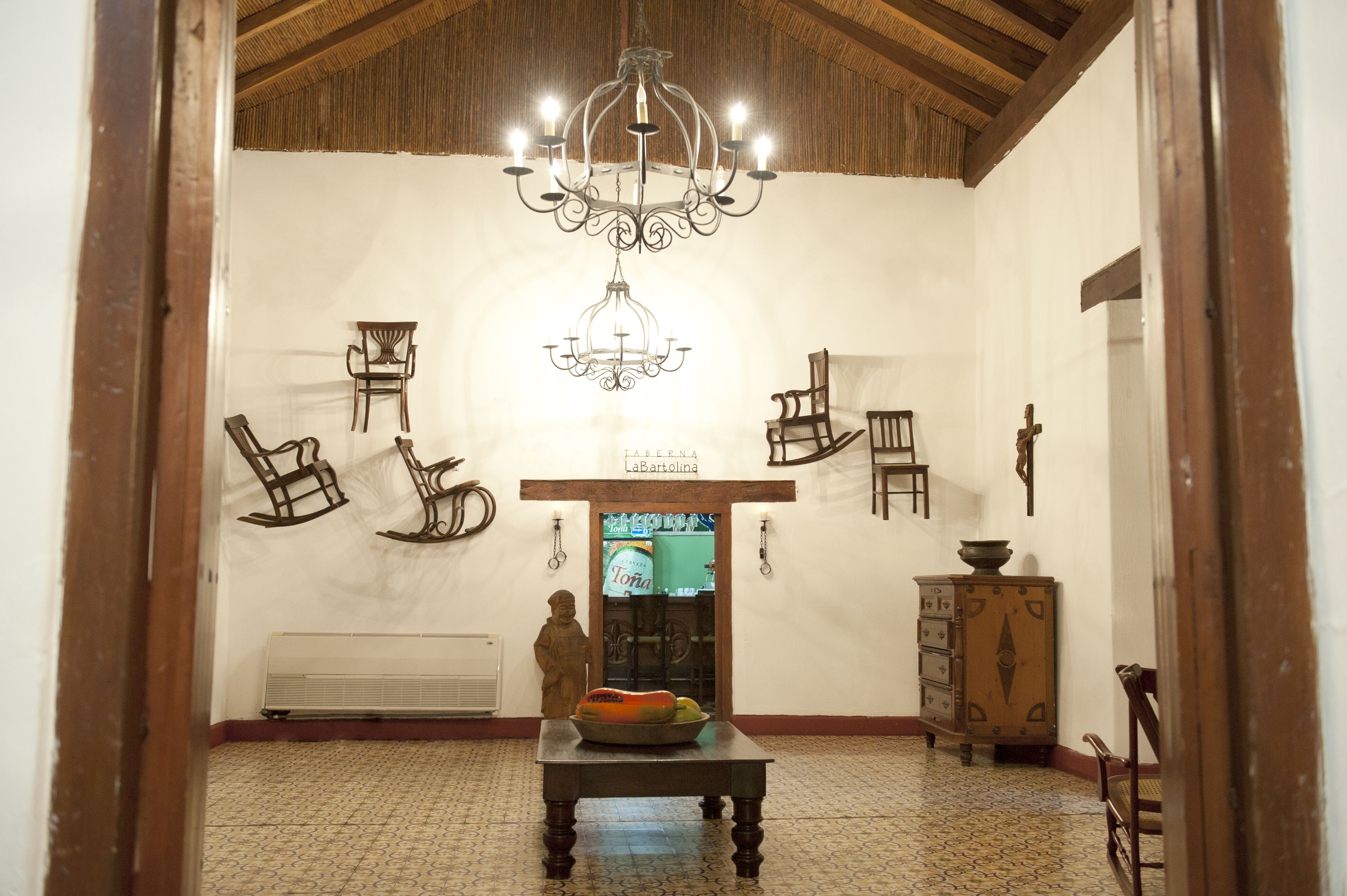 Una habitación con una araña, sillas de madera, una mesa con un cuenco, una estatua y un armario.