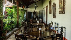 Uma área de refeições com mesas e cadeiras de madeira, piso xadrez, ventilador de teto e uma parede com obras de arte emolduradas.