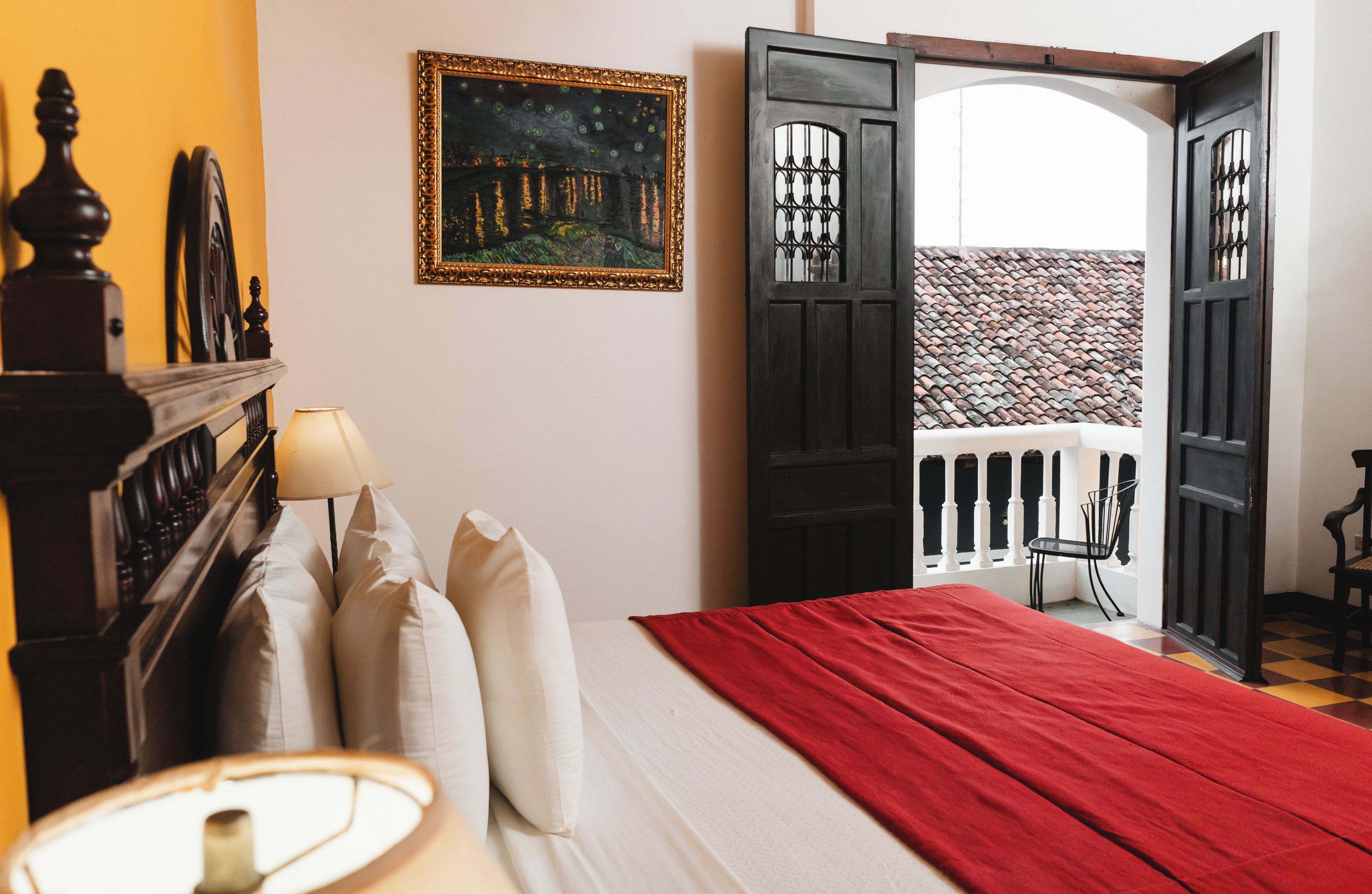 A hotel room with a bed, a nightstand, a lamp, a painting, and an open door leading to a balcony with a view of a tiled roof.