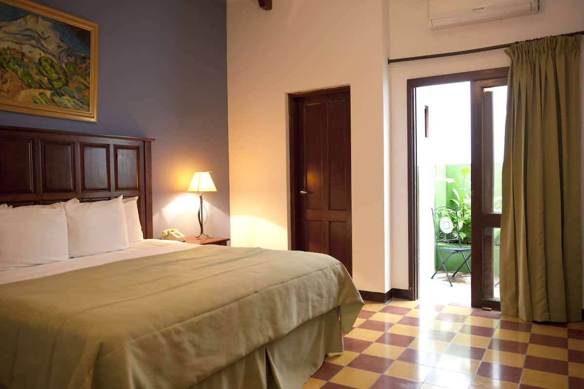 A hotel room with a bed, a nightstand, a lamp, a door, and a view of the outside.