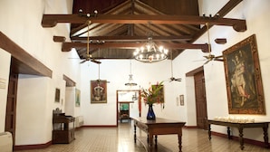 Um corredor amplo com teto alto, vigas de madeira, um candelabro, ventiladores de teto e uma mesa com um vaso de flores.