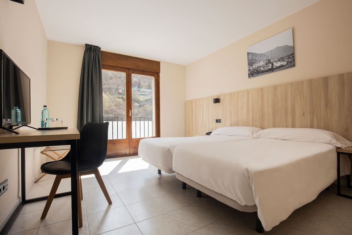 A hotel room with two beds, a desk with a chair, a television, and a window with a view of mountains.