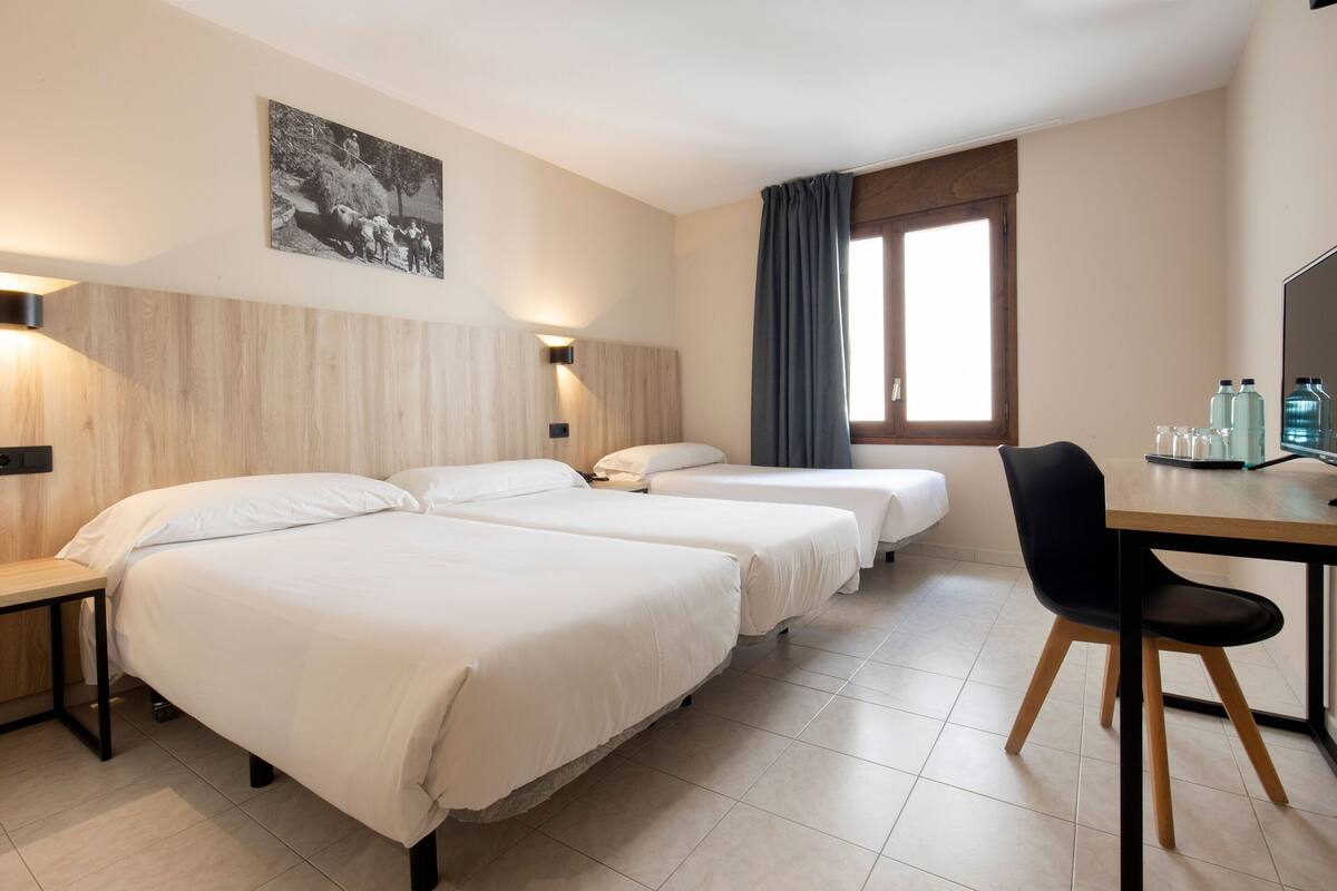 A hotel room with two beds, a desk with a chair, a window with curtains, and a wall-mounted picture.