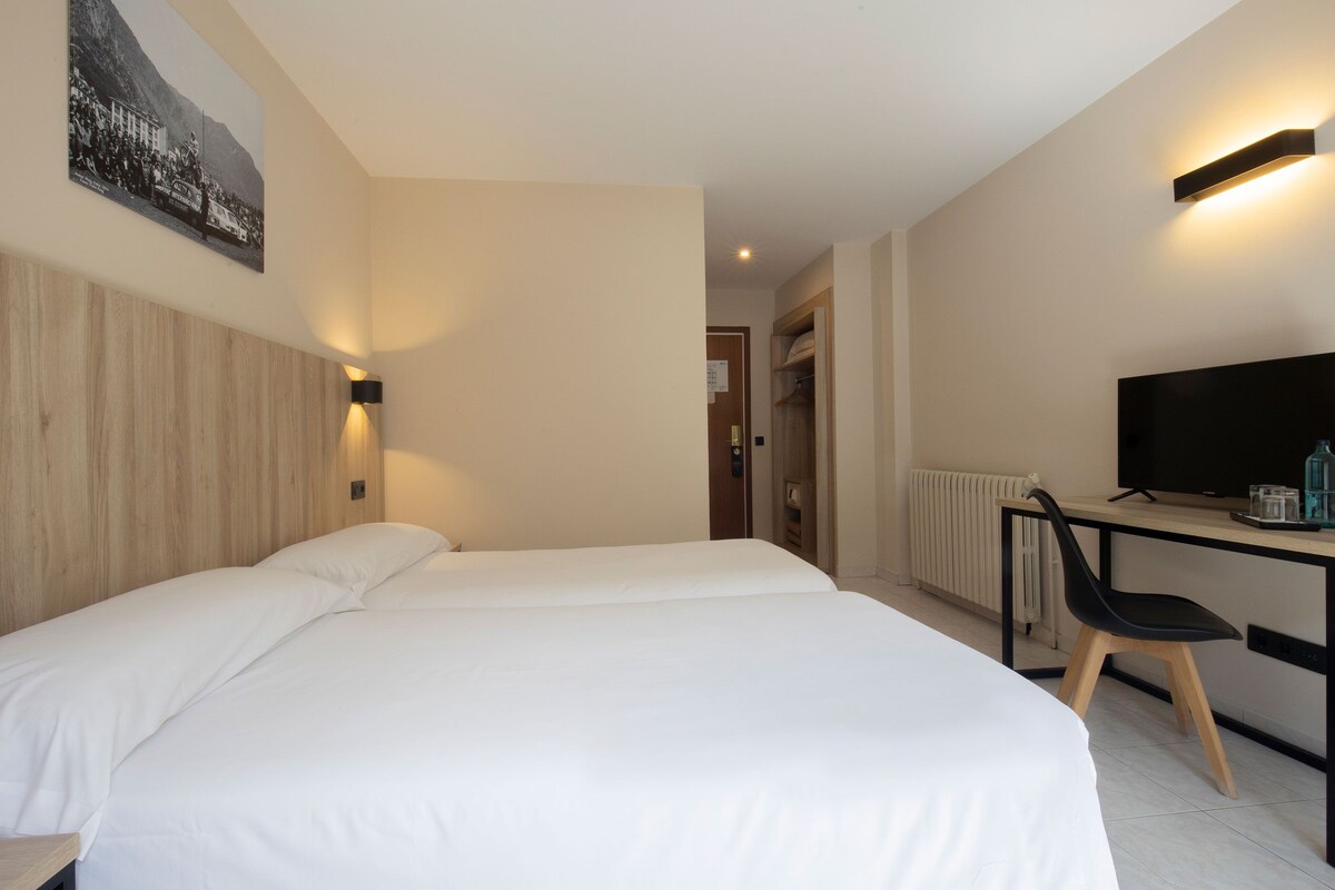 Quarto de hotel com cama, escrivaninha com cadeira, televisão e luminária na parede.