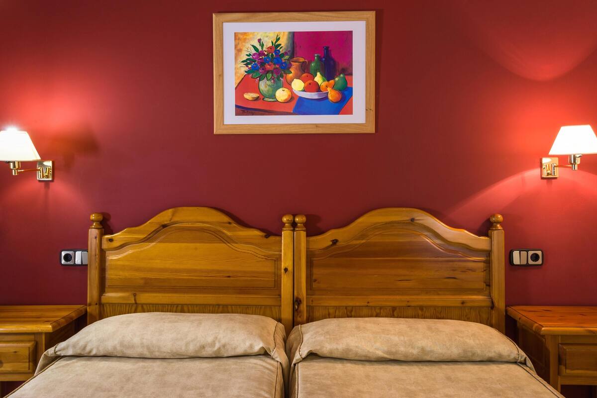 A double bed with a wooden headboard, two bedside tables with lamps, a painting on the wall, and a red wall.