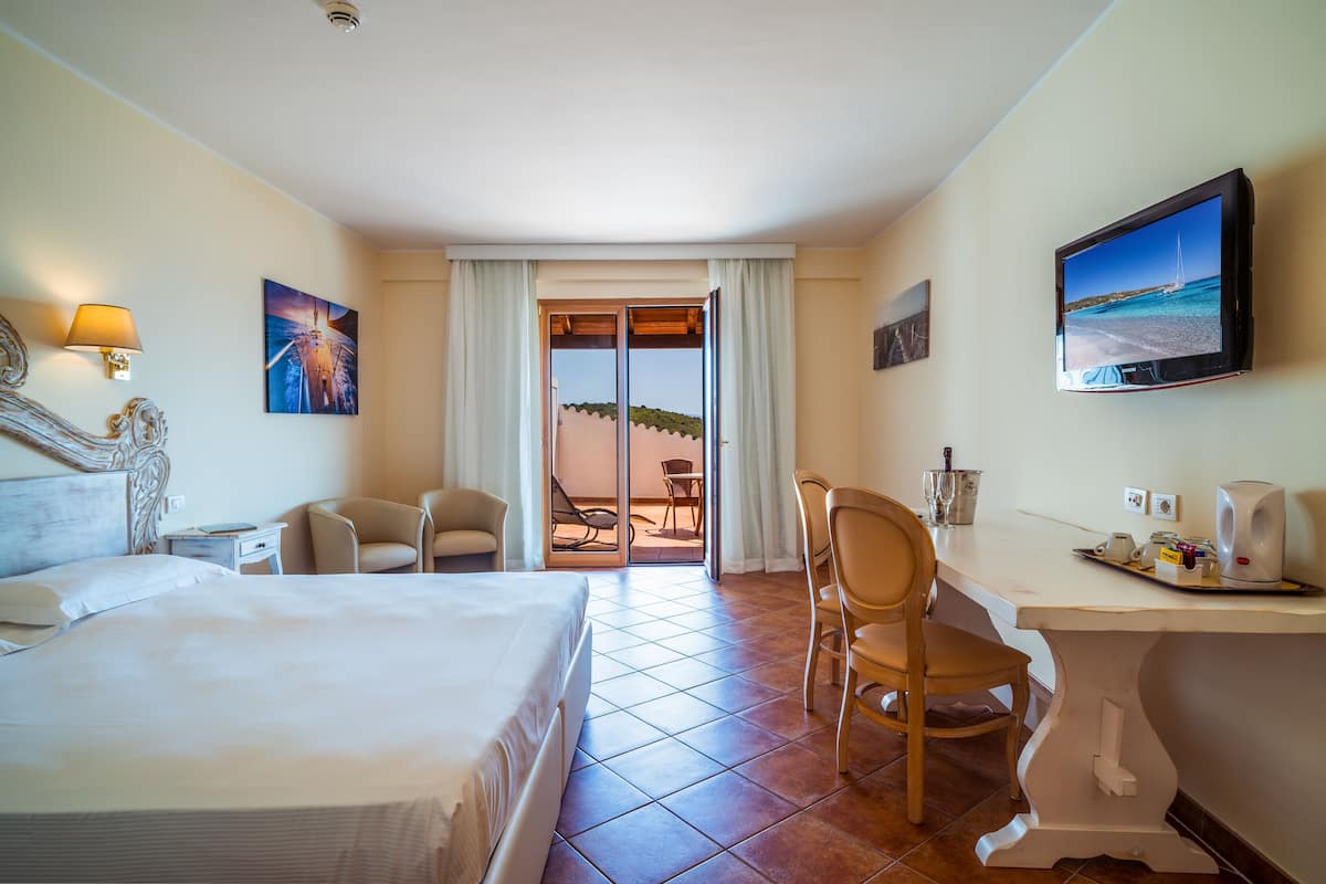 A hotel room with a bed, a desk, a chair, a television, and a view of the beach through the door.