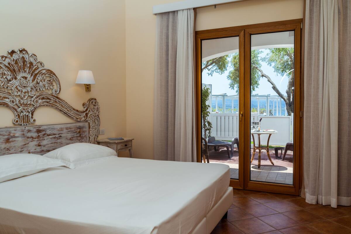 A hotel room with a large bed, a nightstand, a lamp, and a sliding glass door leading to an outdoor patio with a table and chairs.