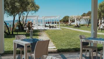 A covered outdoor dining area with tables and chairs, a pathway leading to a pool, and a view of the sea.