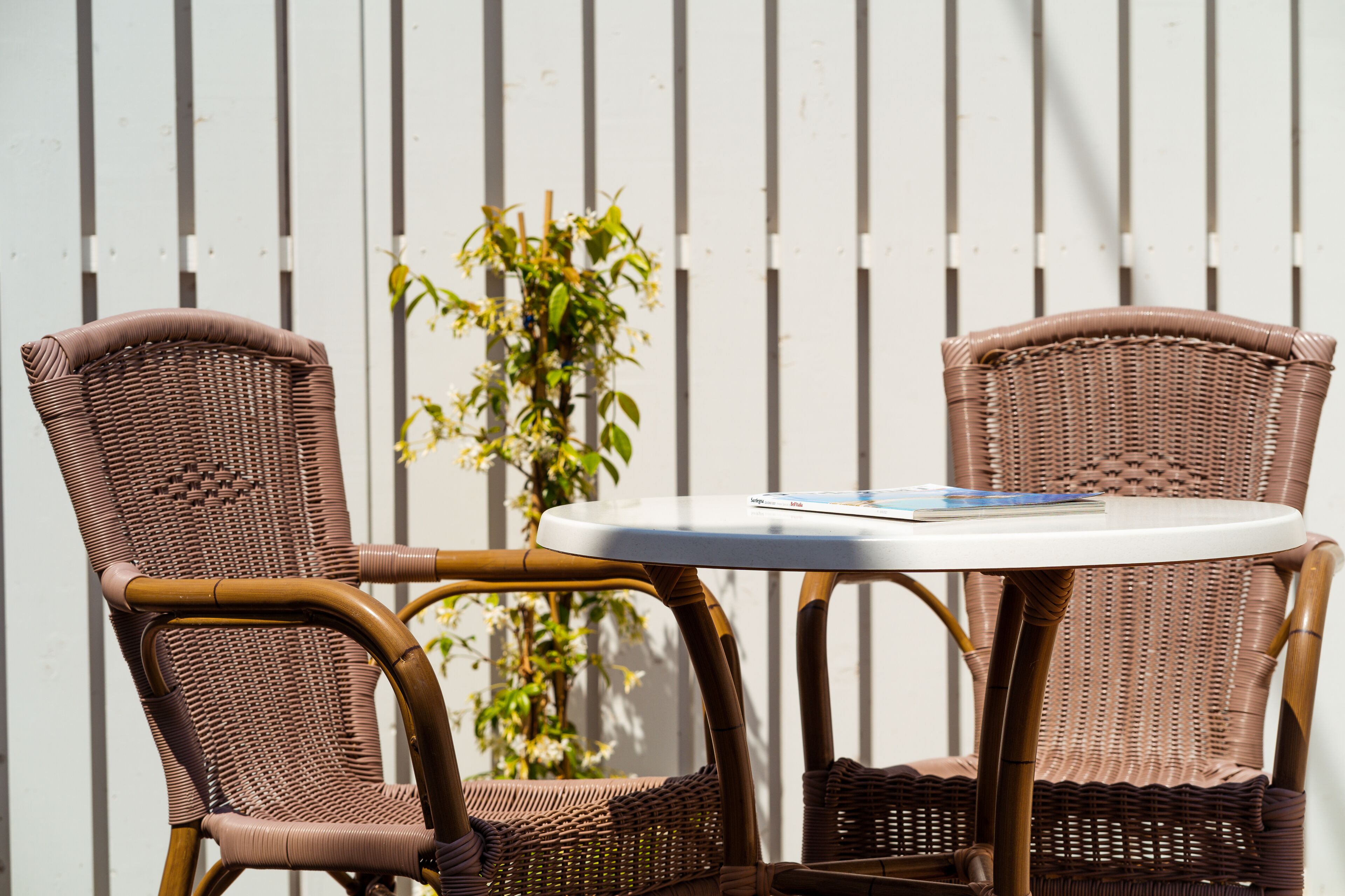 Two wicker chairs with a small round table between them, set against a white fence with vertical lines.