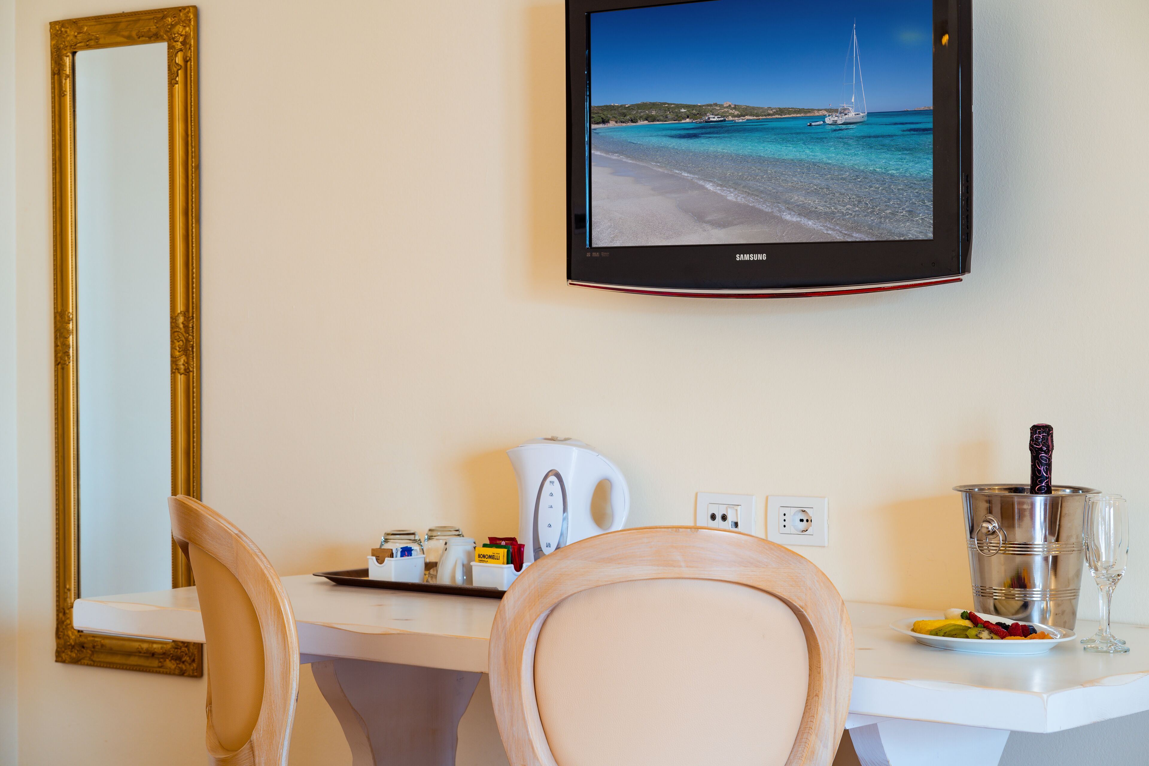 Habitación de hotel con una televisión mostrando una escena de playa, un escritorio blanco con una tetera, un plato de fruta y una copa de vino.