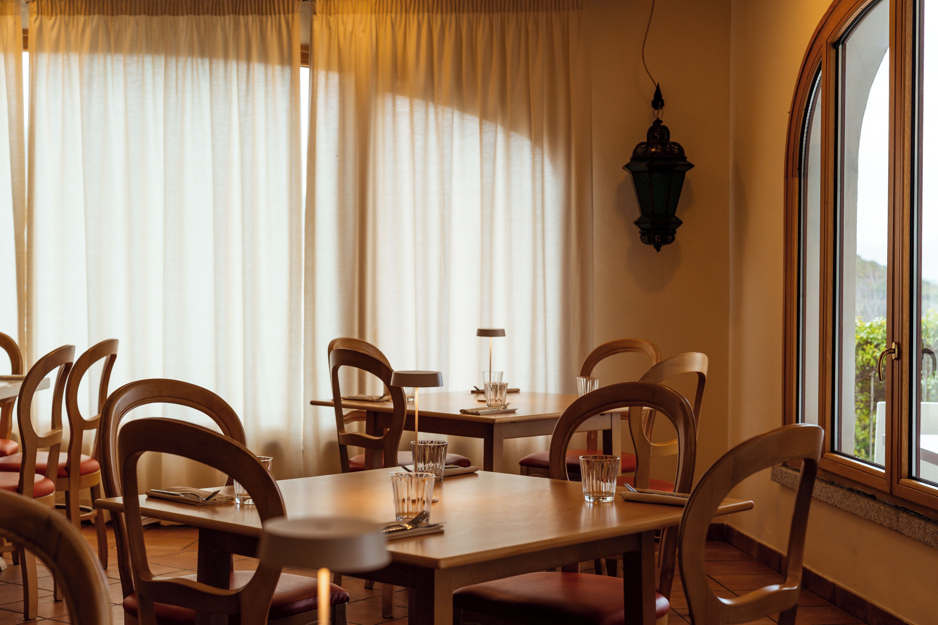 A dining area with wooden tables and chairs, a hanging lantern, and a window with a view.