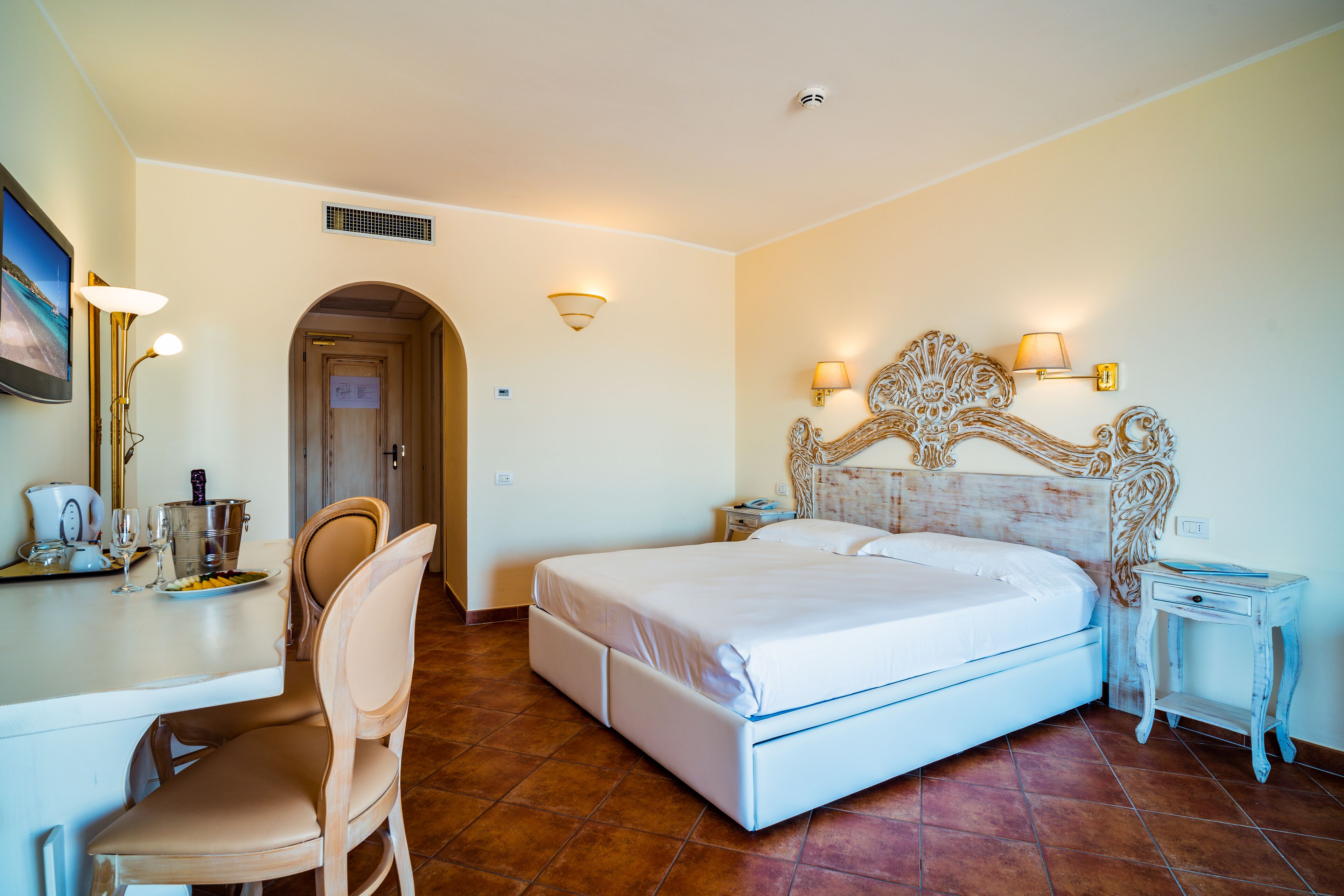 A hotel room with a bed, a desk with a chair, a nightstand, and a wall-mounted lamp.