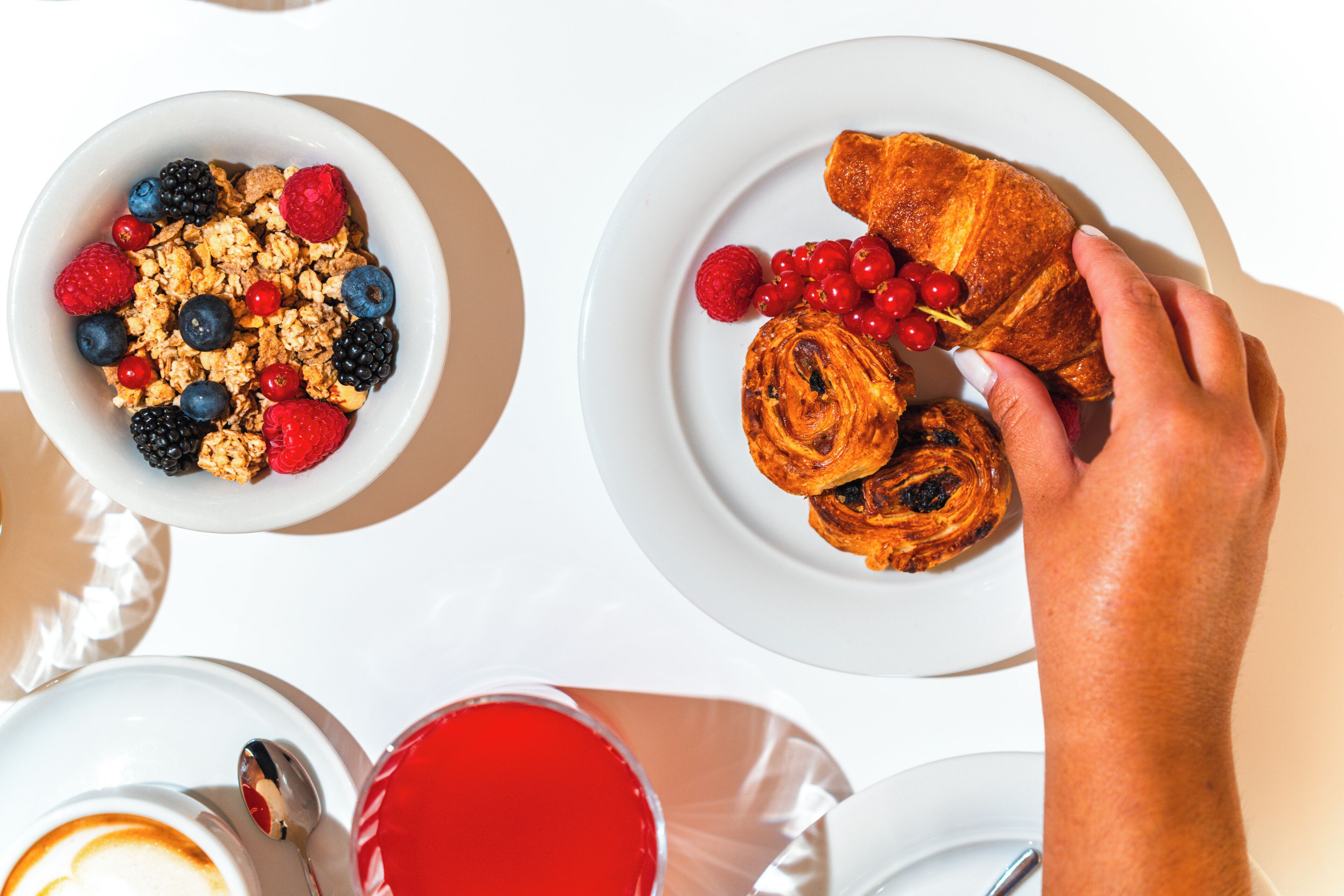 A breakfast setting with a bowl of granola and berries, a plate with croissants and berries, and a hand reaching for a croissant.