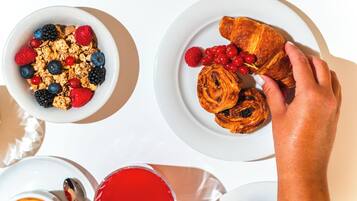 Une table mise pour le petit-déjeuner avec un bol de granola et des fruits rouges, une assiette avec des croissants et des fruits rouges, et une main qui tend la main vers un croissant.