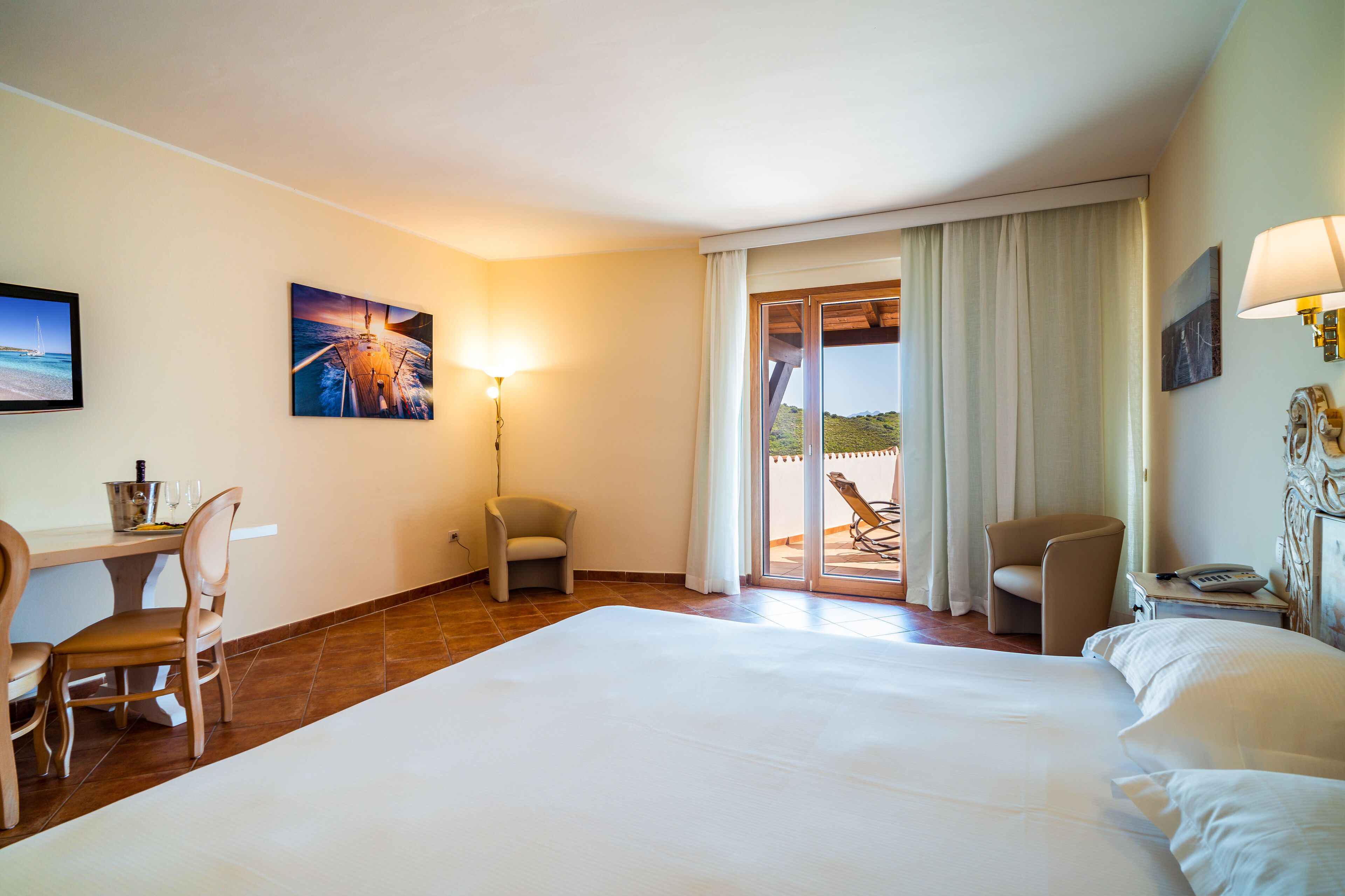 A hotel room with a large bed, a desk with a chair, a small table with a lamp, and a view of the outdoors through a sliding door.