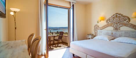 A hotel room with a bed, a desk, chairs, a nightstand, and a view of the sea.