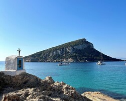 Costa rocosa con un pequeño edificio que tiene una cruz en la parte superior, barcos en el agua y una colina destacada al fondo.
