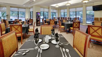 A dining area with multiple tables set for service, large windows, and a television mounted on the wall.