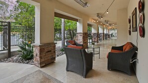 A covered outdoor seating area with wicker furniture, stone pillars, and large windows overlooking greenery.