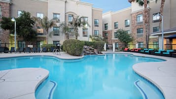 A swimming pool with a clear blue water, surrounded by lounge chairs and a building with multiple windows and balconies.