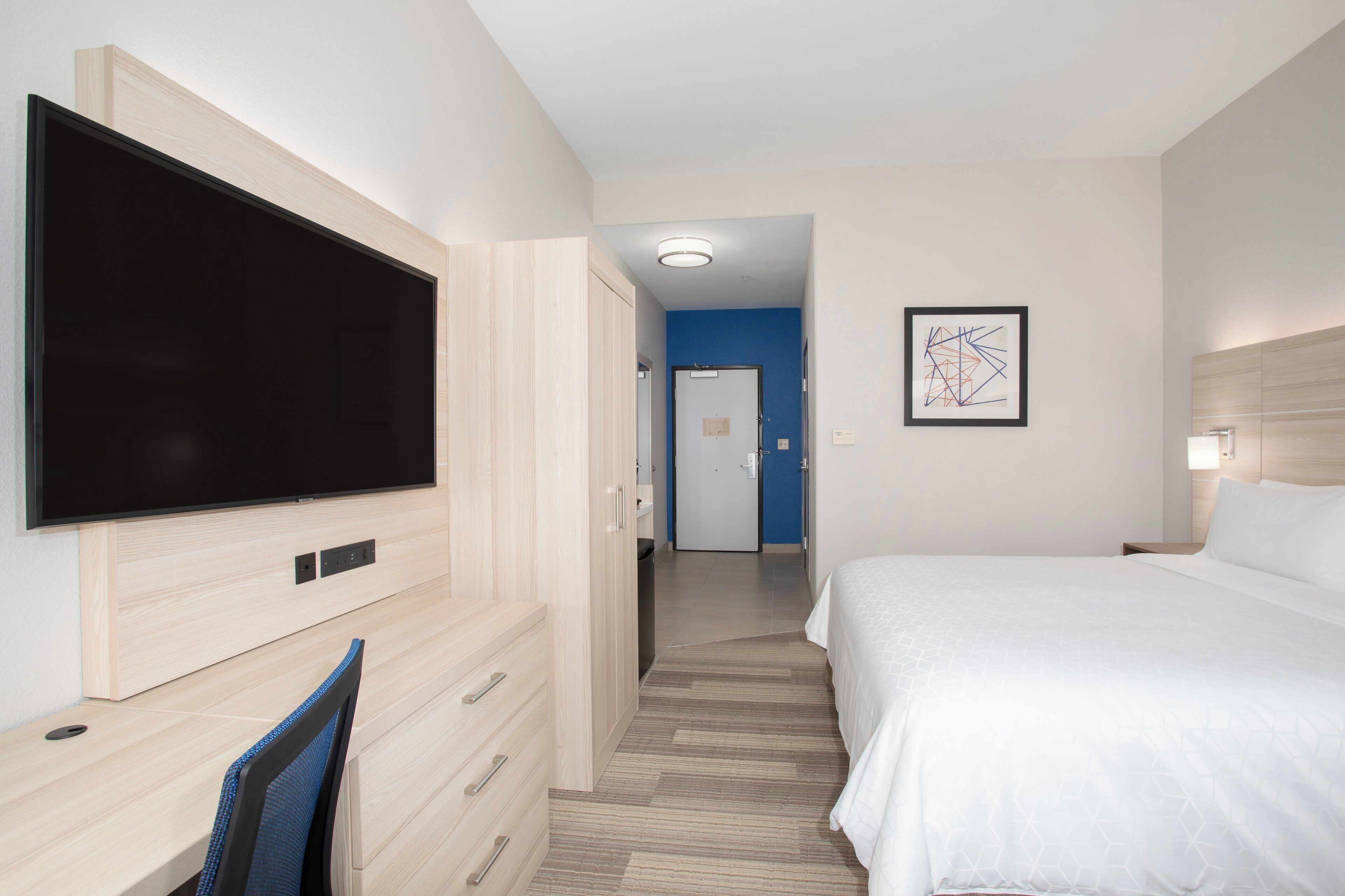 Habitación de hotel con un televisor de pantalla plana grande montado en la pared, una cama con ropa blanca, un escritorio con silla y una obra de arte abstracta enmarcada en la pared.