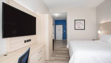Habitación de hotel con un televisor de pantalla plana grande montado en la pared, una cama con ropa blanca, un escritorio con silla y una obra de arte abstracta enmarcada en la pared.