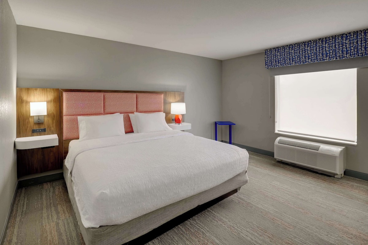 Habitación de hotel con una cama grande, dos mesitas de noche con lámparas, una ventana con vistas y un aire acondicionado instalado en la pared.