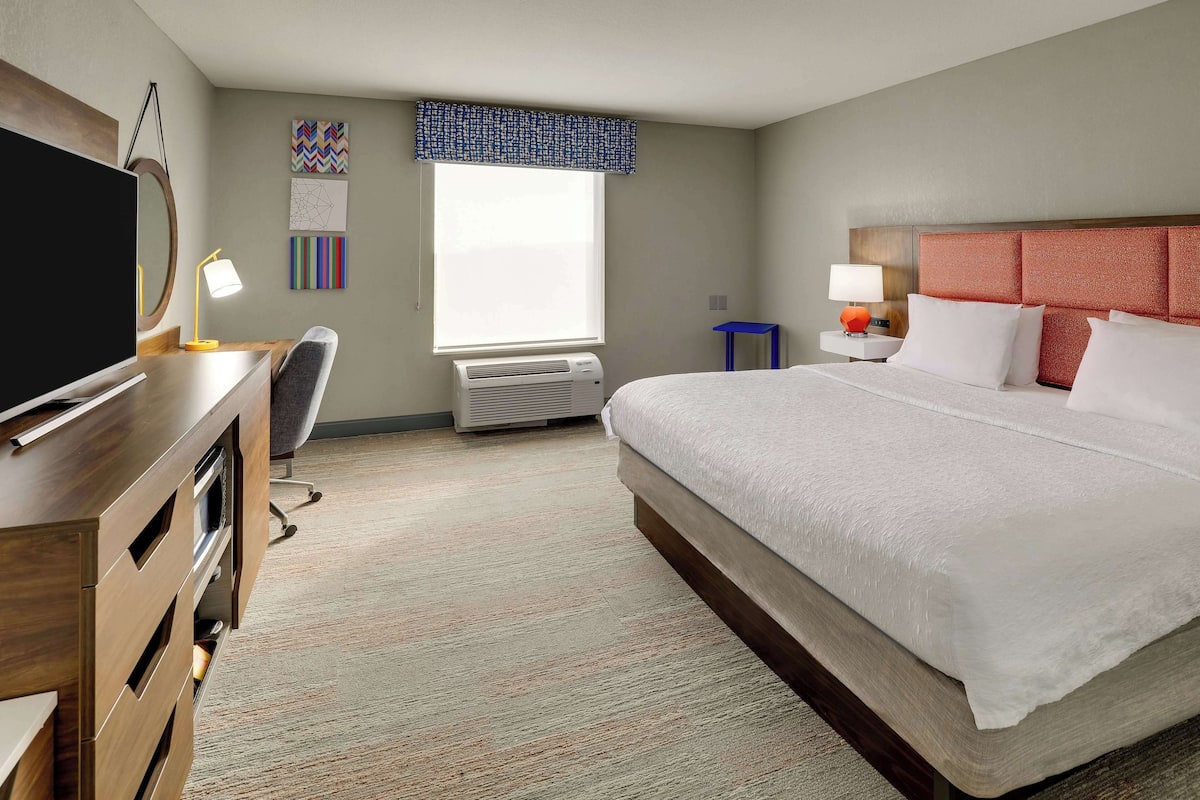 Habitación de hotel con una cama grande, un escritorio con televisor, una silla, una lámpara de escritorio y un ventanal con cortinilla azul.
