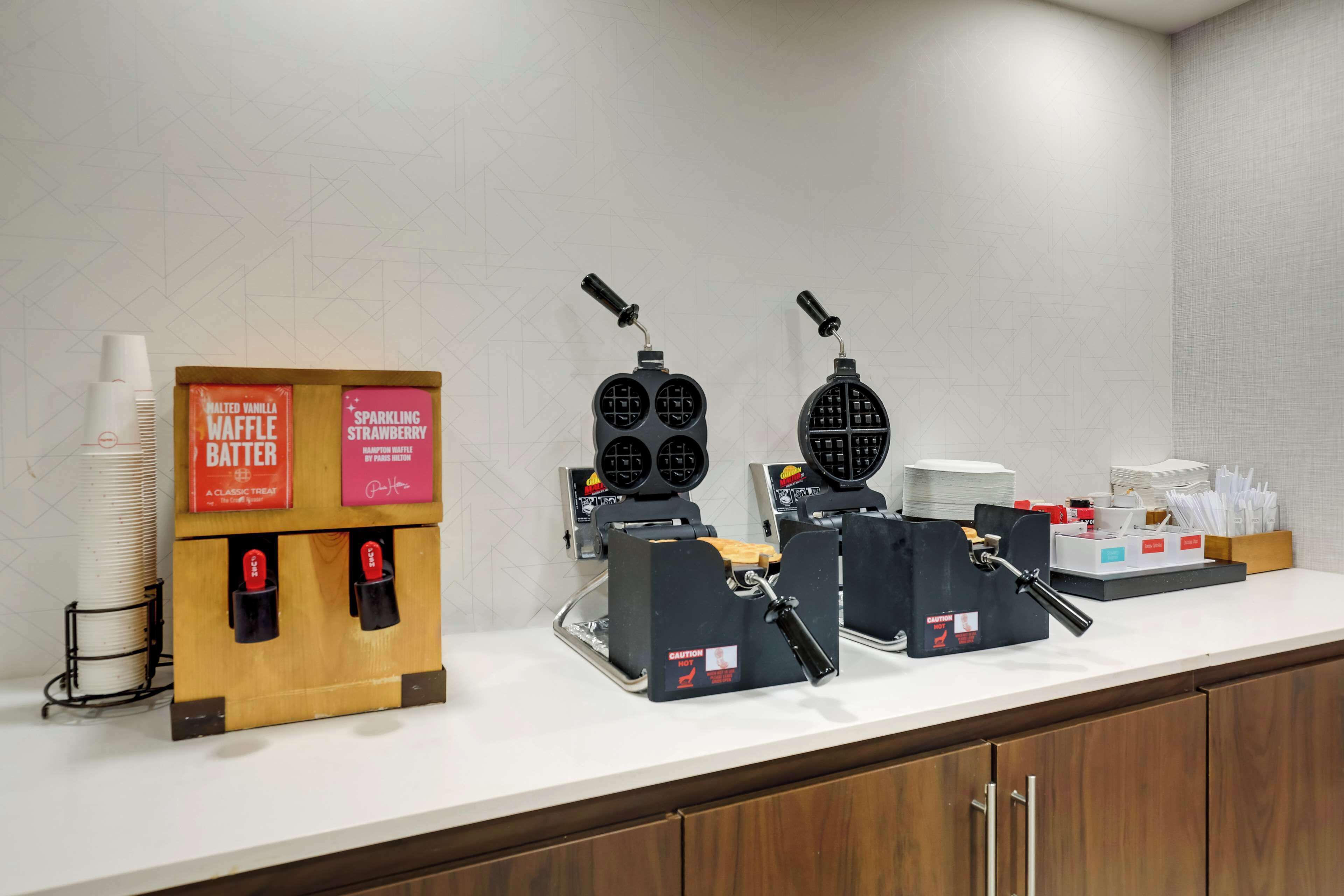 A breakfast station with waffle makers, waffle batter boxes, and disposable cups.