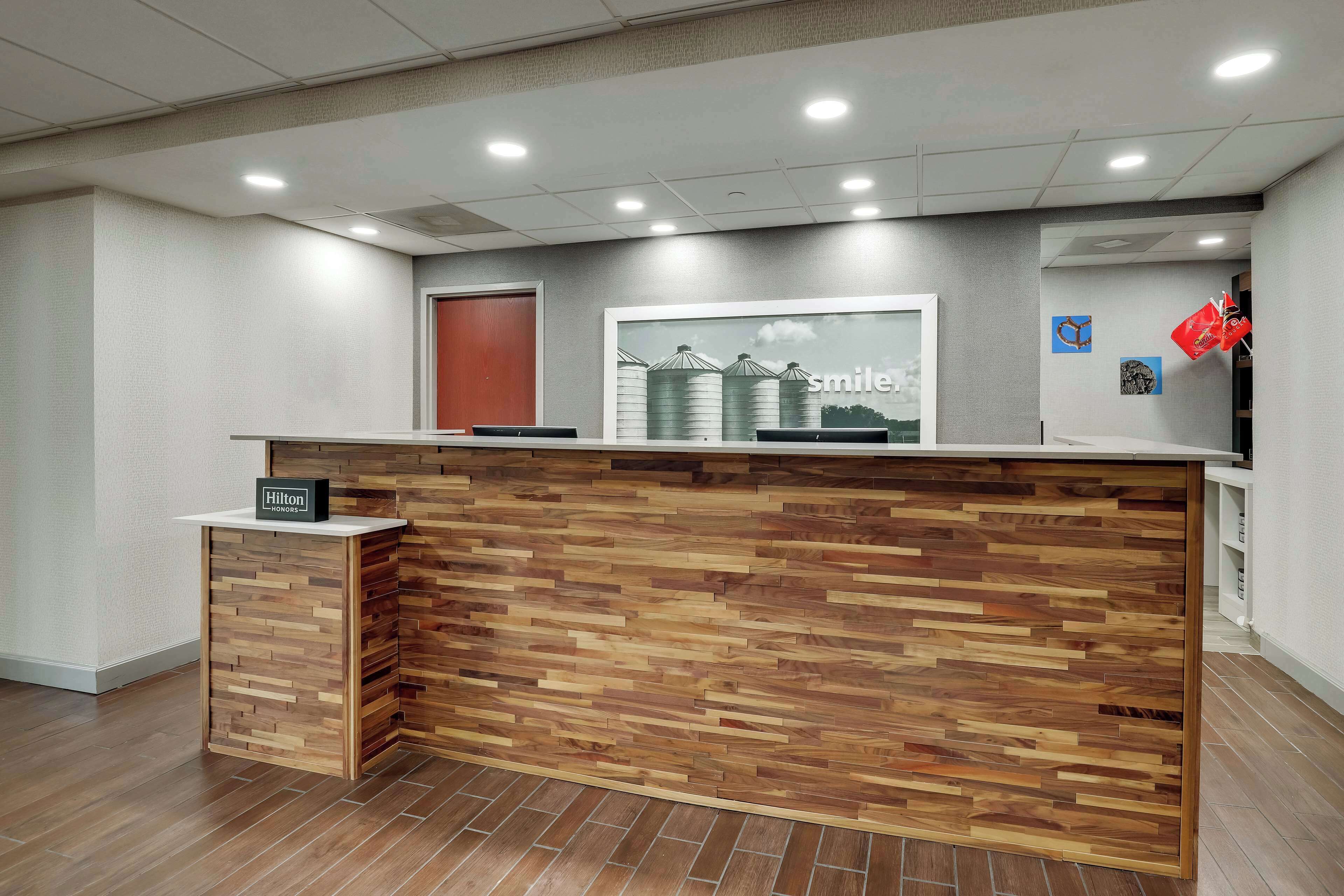 A hotel reception desk with a wooden panel design, a sign indicating Hilton Honors, and a framed picture of a farm on the wall.