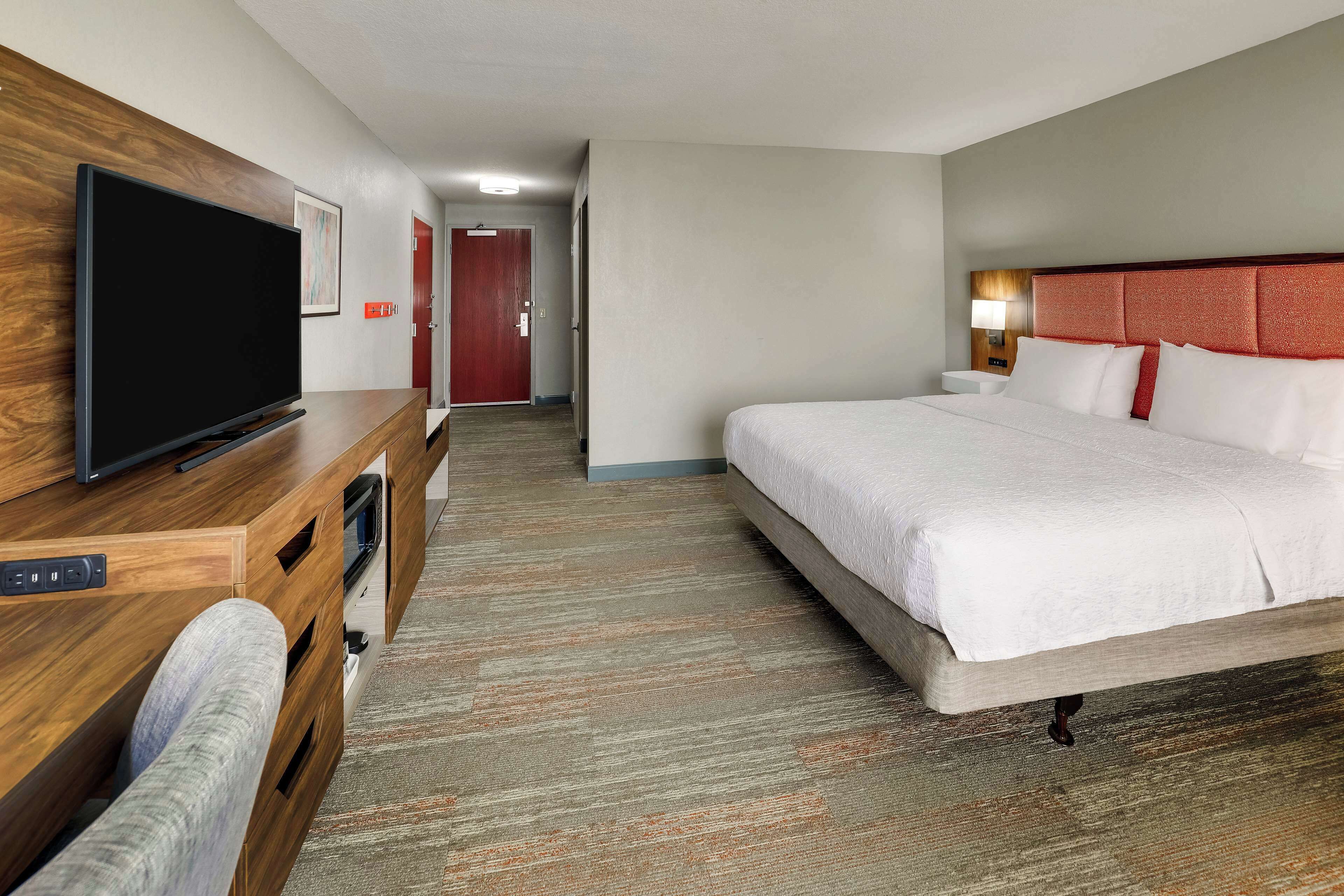 Habitación de hotel con una cama grande, un televisor sobre un aparador de madera, piso alfombrado y un pasillo que conduce a una puerta.