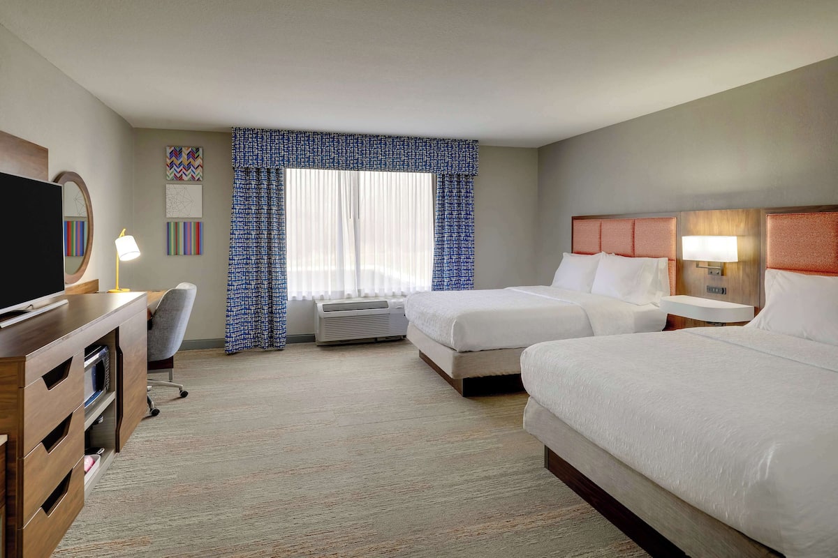 Una habitación de hotel con dos camas, un escritorio con televisor, una silla y un ventanal con cortinas.