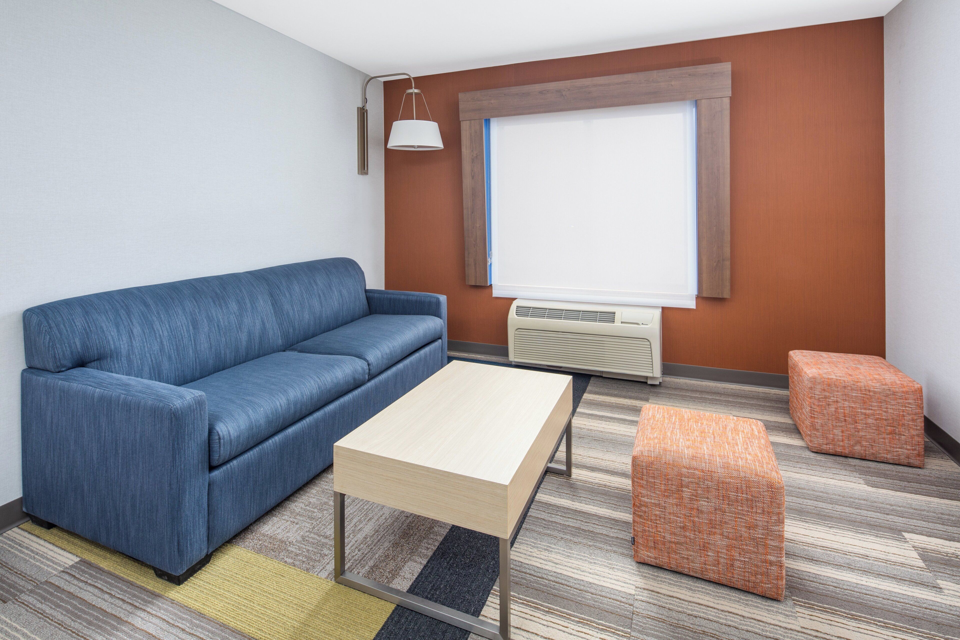 Una habitación con un sofá azul, una mesa de centro de madera, un pequeño taburete naranja, una ventana con persianas y un aire acondicionado instalado en la pared.