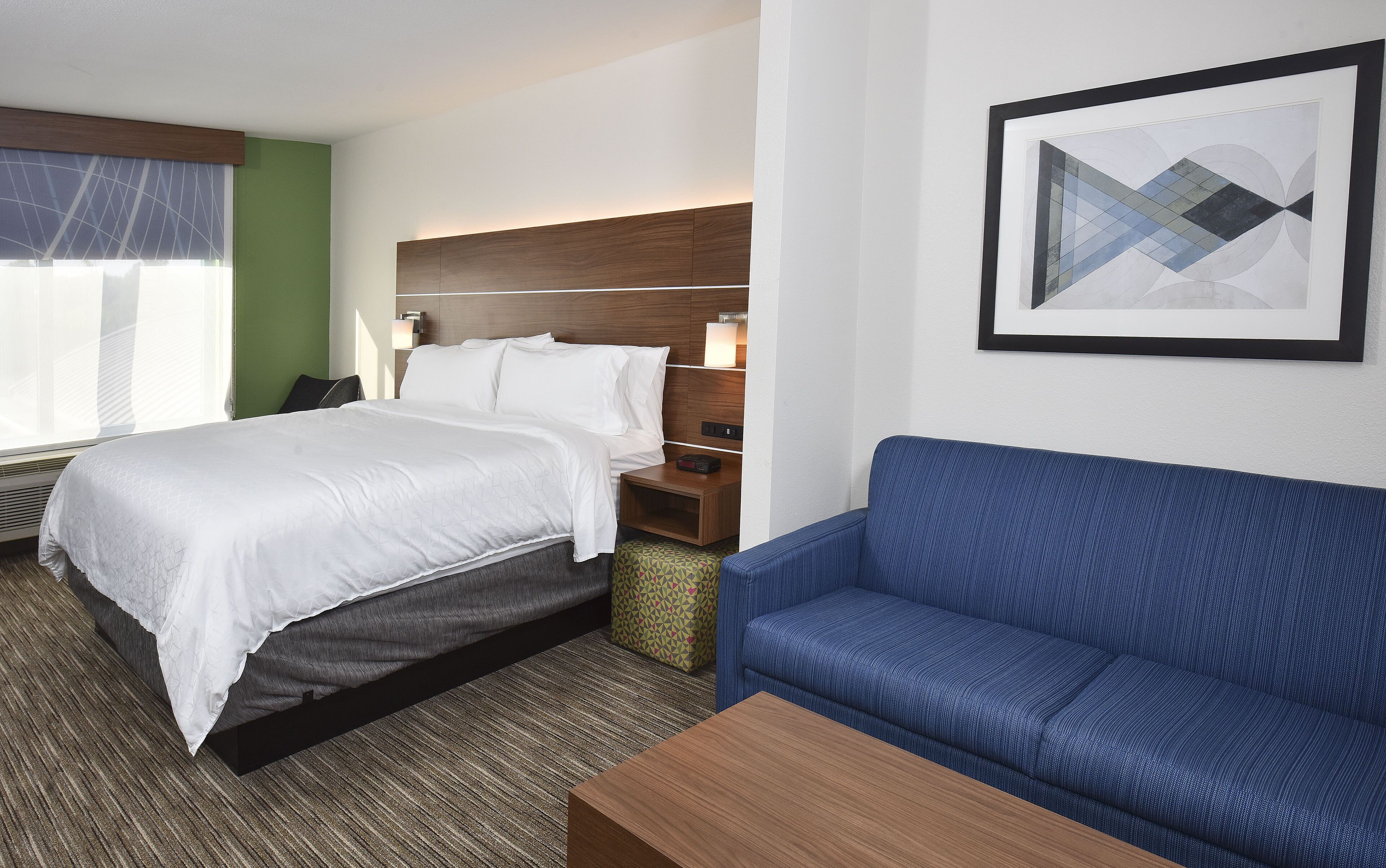 A hotel room with a bed, a blue sofa, a wooden nightstand, and a framed abstract artwork on the wall.