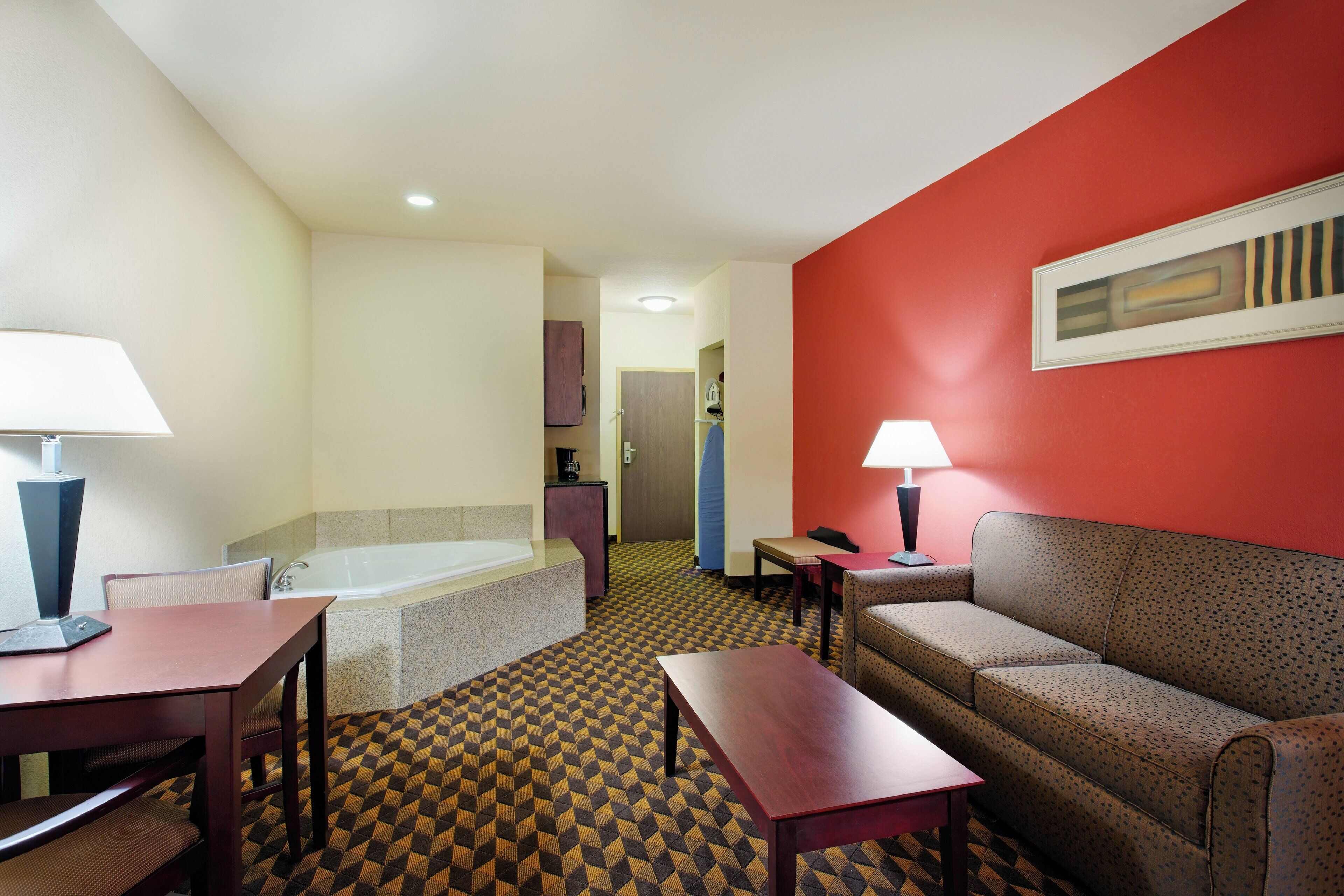 A hotel room with a bed, a sitting area with a sofa and a table, a small table with a lamp, and a bathroom with a bathtub and a shower.