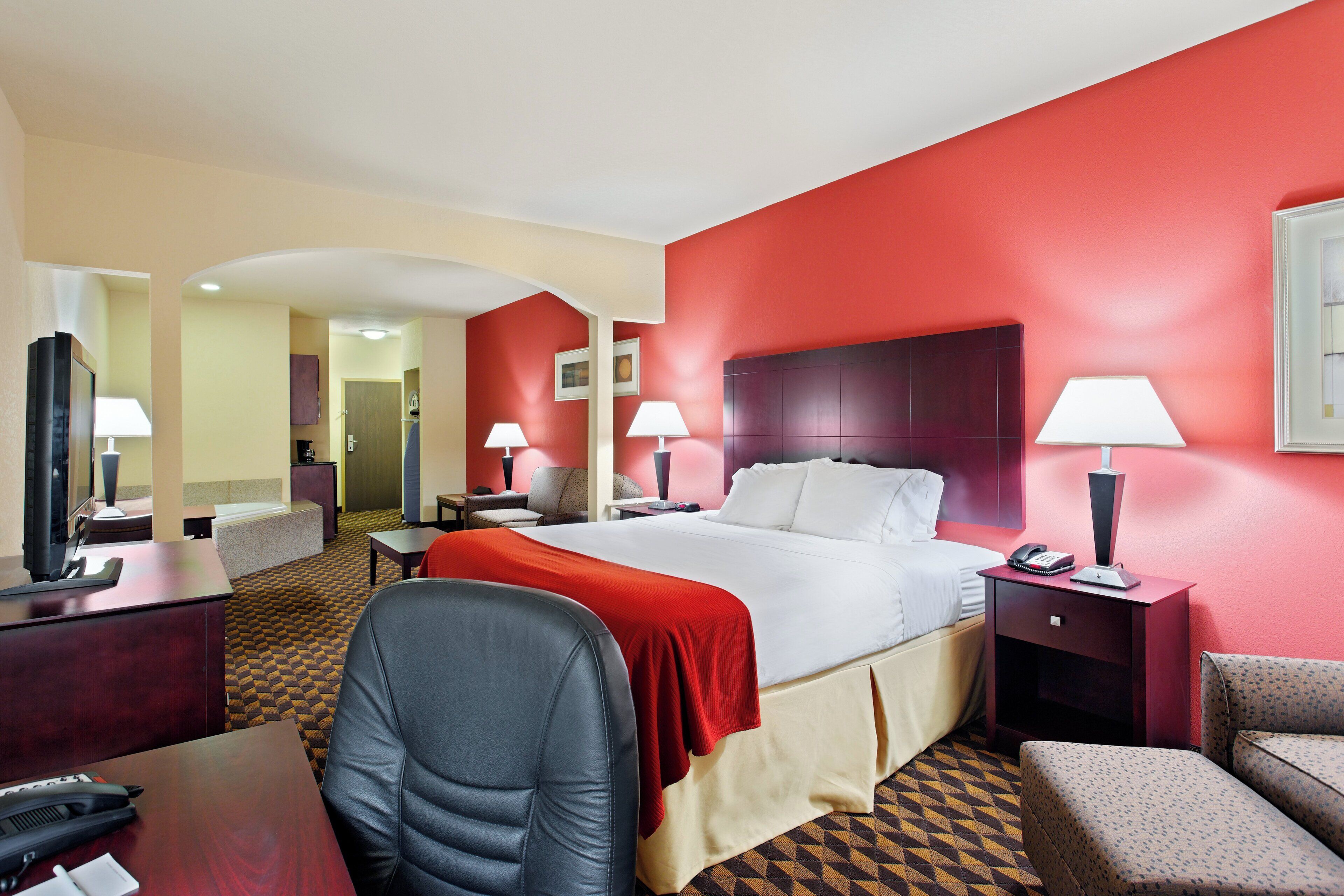 A hotel room with a large bed, two bedside tables with lamps, a desk with a telephone, a chair, and a patterned carpet.