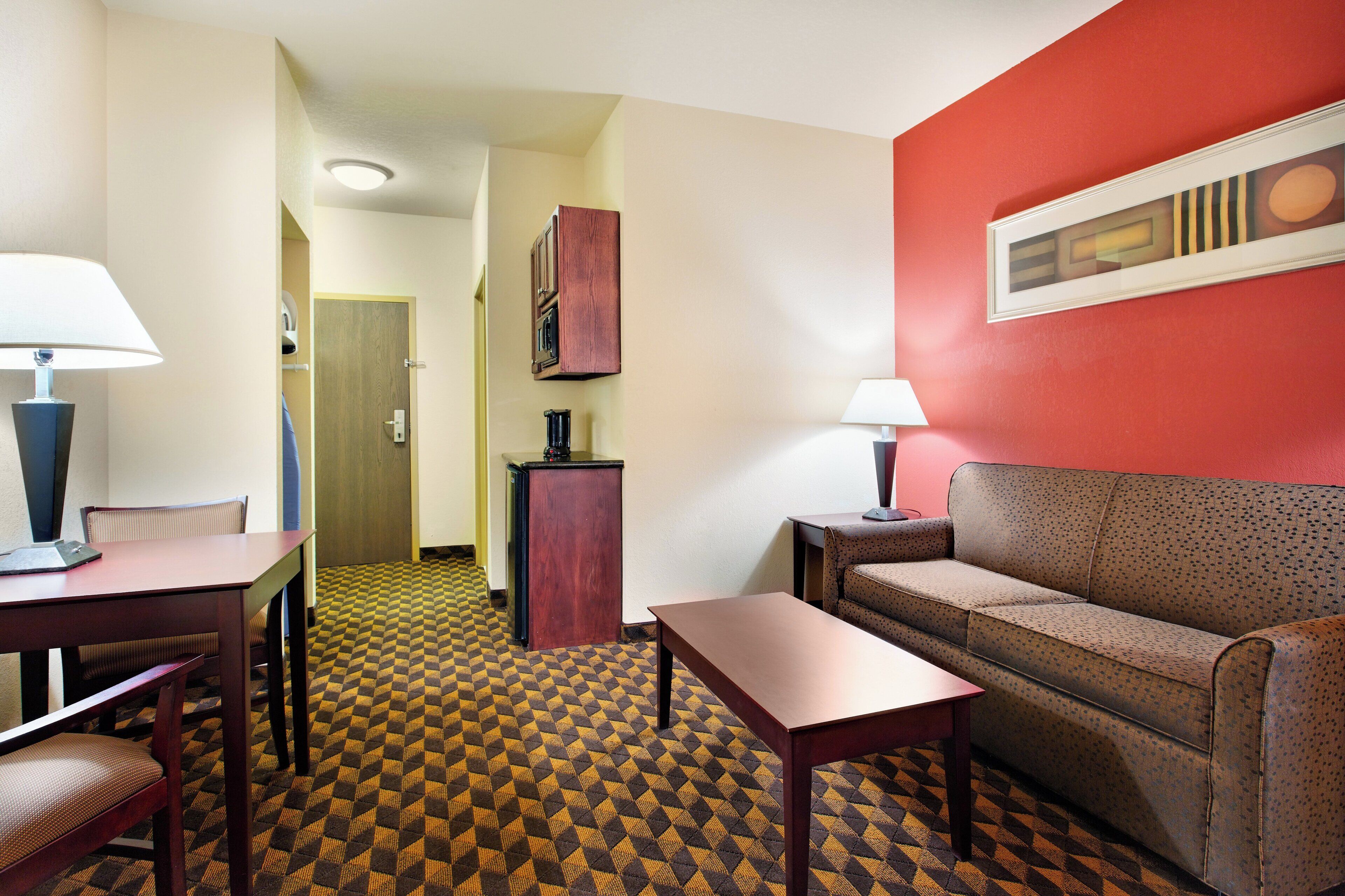 Une chambre d’hôtel avec une moquette à motifs, un canapé, une table, un bureau avec une lampe et un placard.