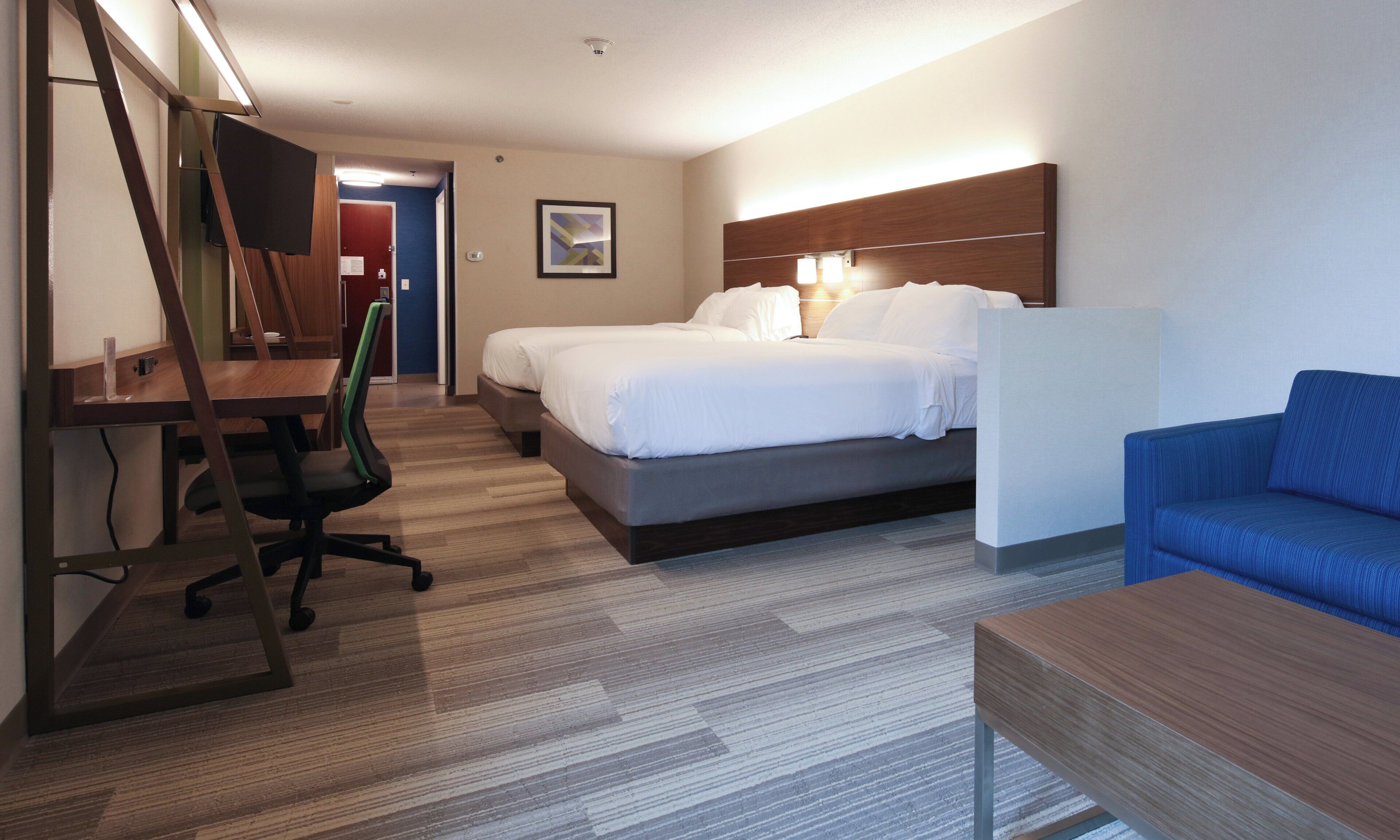 Una habitación de hotel con escritorio, dos camas, un sofá azul y un cabecero de madera.