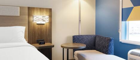 A hotel room with a bed, a small table, a chair, a lamp, and a window with blinds.
