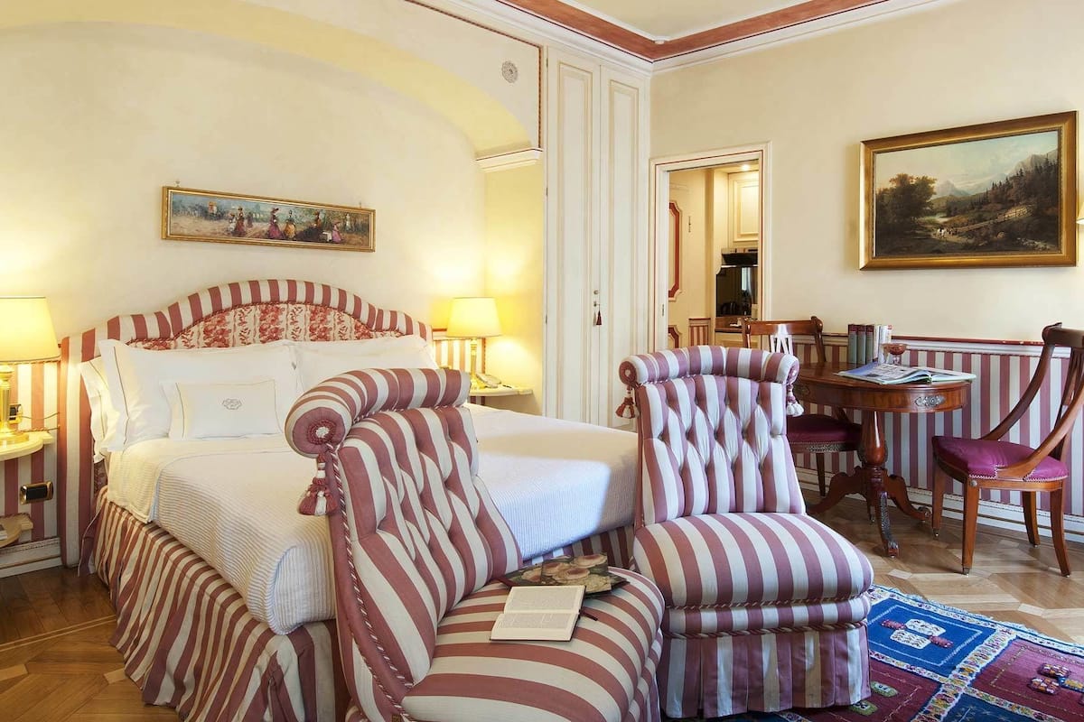Een hotelkamer met een groot bed, twee gestreepte fauteuils, een tafeltje met een lamp en een schilderij aan de muur.