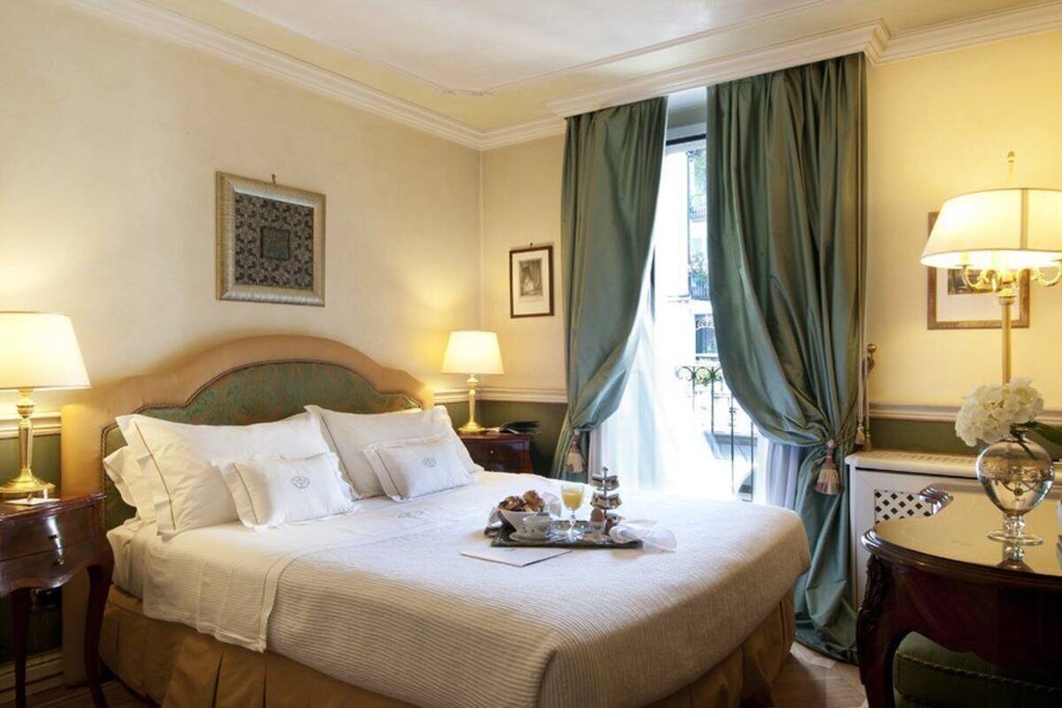 Een hotelkamer met een groot bed, twee nachtkastjes met lampen, een kleine tafel met een vaas, en uitzicht door een raam met gordijnen.