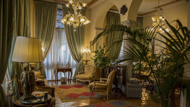 Un salon luxueux, agrémenté de meubles richement décorés, d’un grand lustre et de divers objets décoratifs.