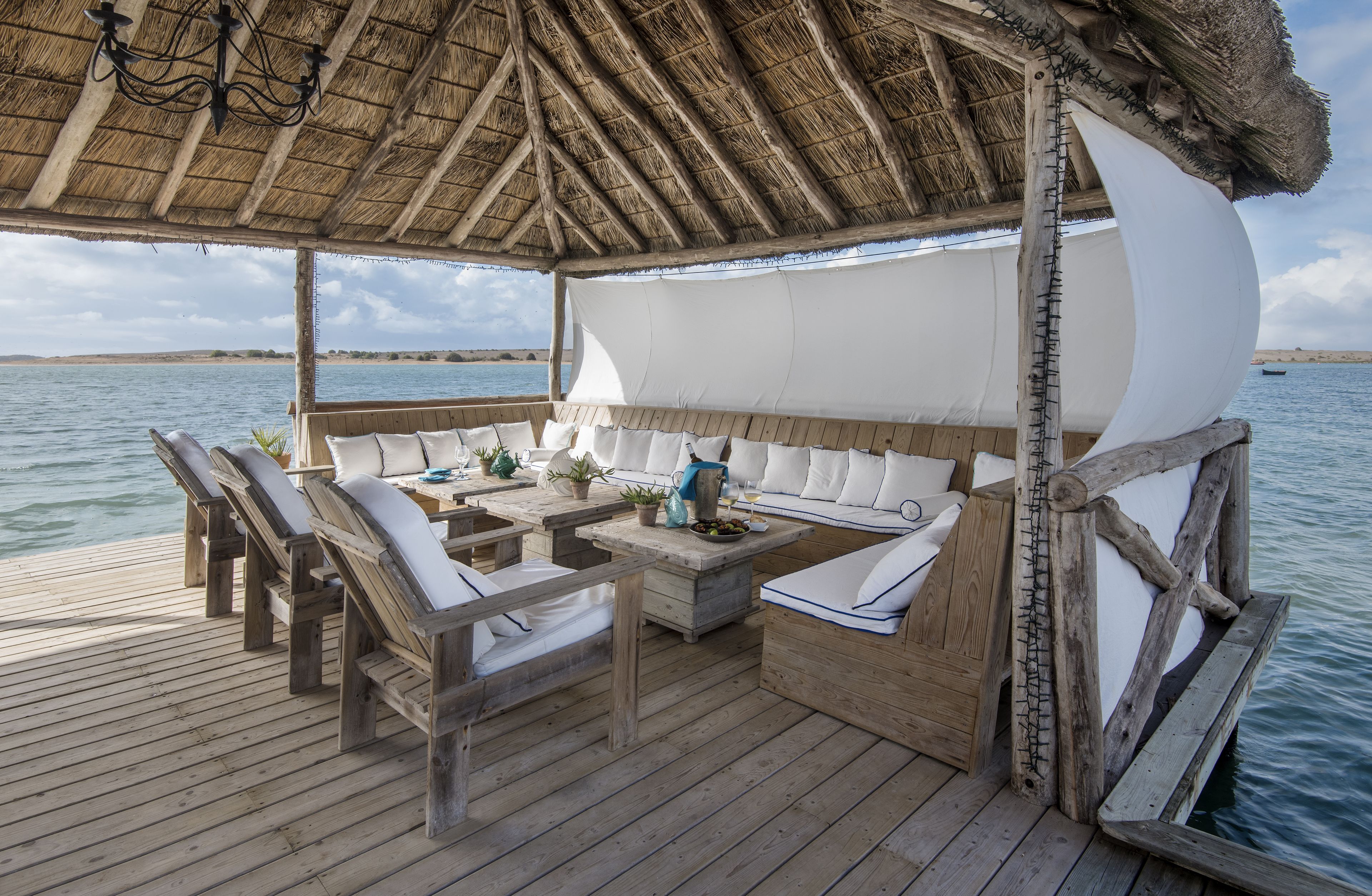 A wooden deck with a thatched roof, white cushioned seating, and a table with drinks and snacks.