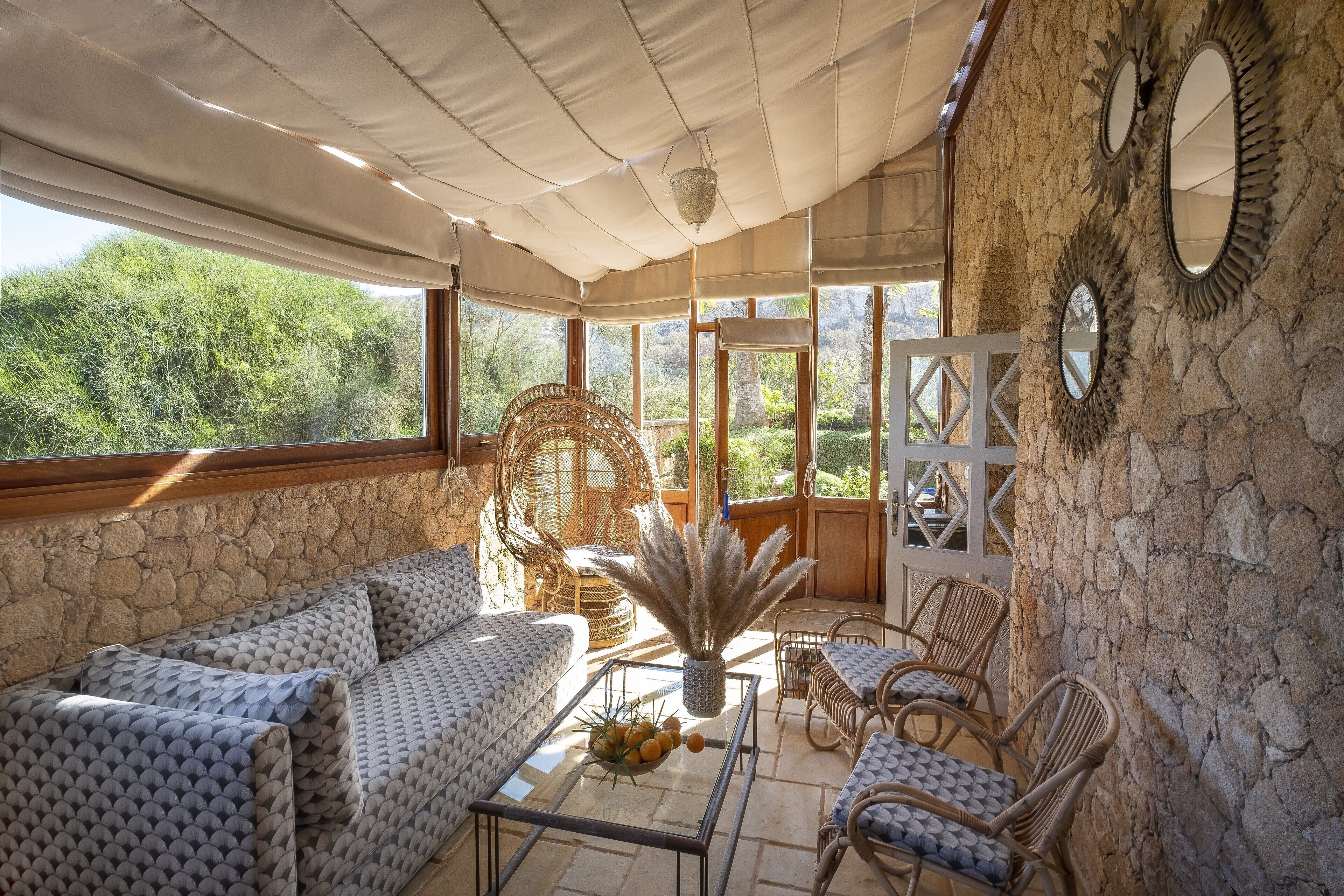 A sunlit patio with stone walls, wicker furniture, and a view of greenery.