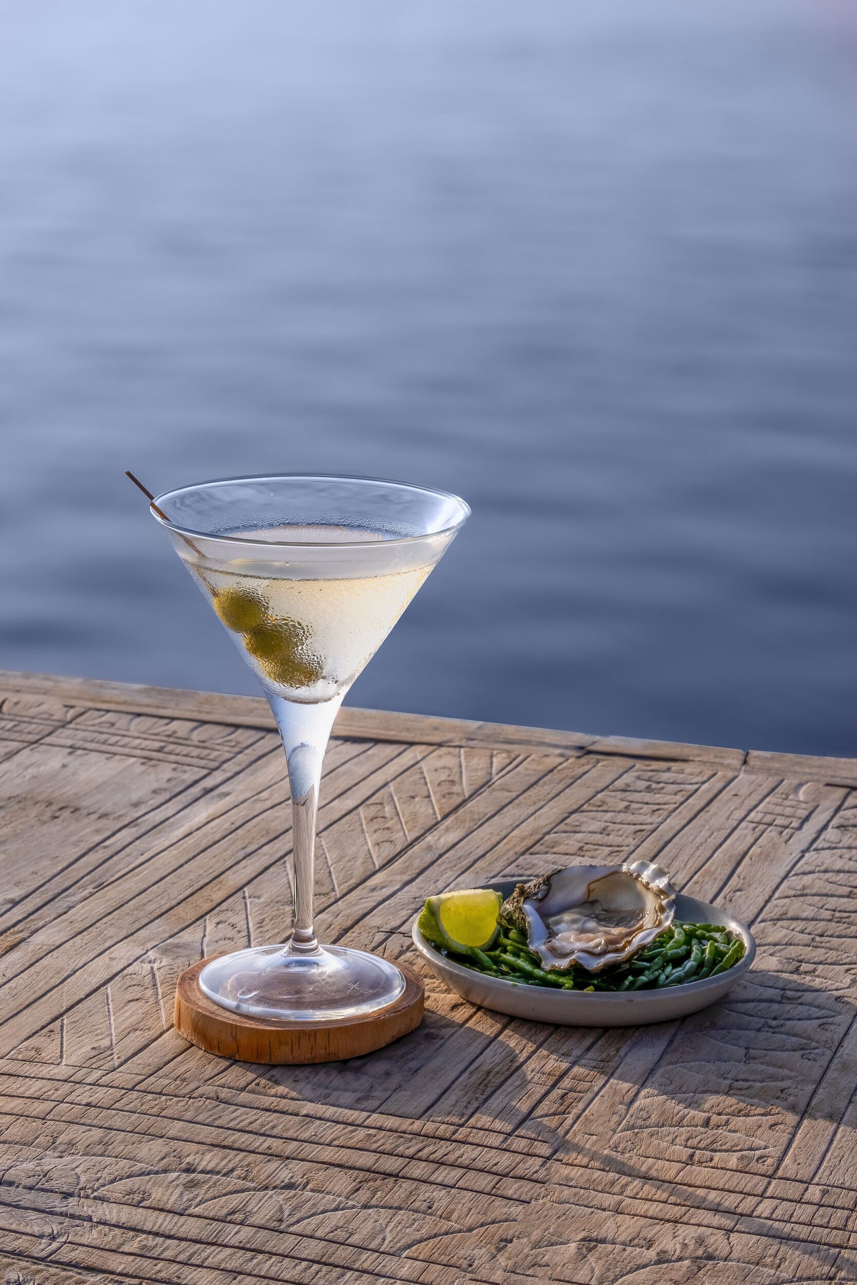 A martini glass on a wooden surface with a plate of oysters and lemon beside it.