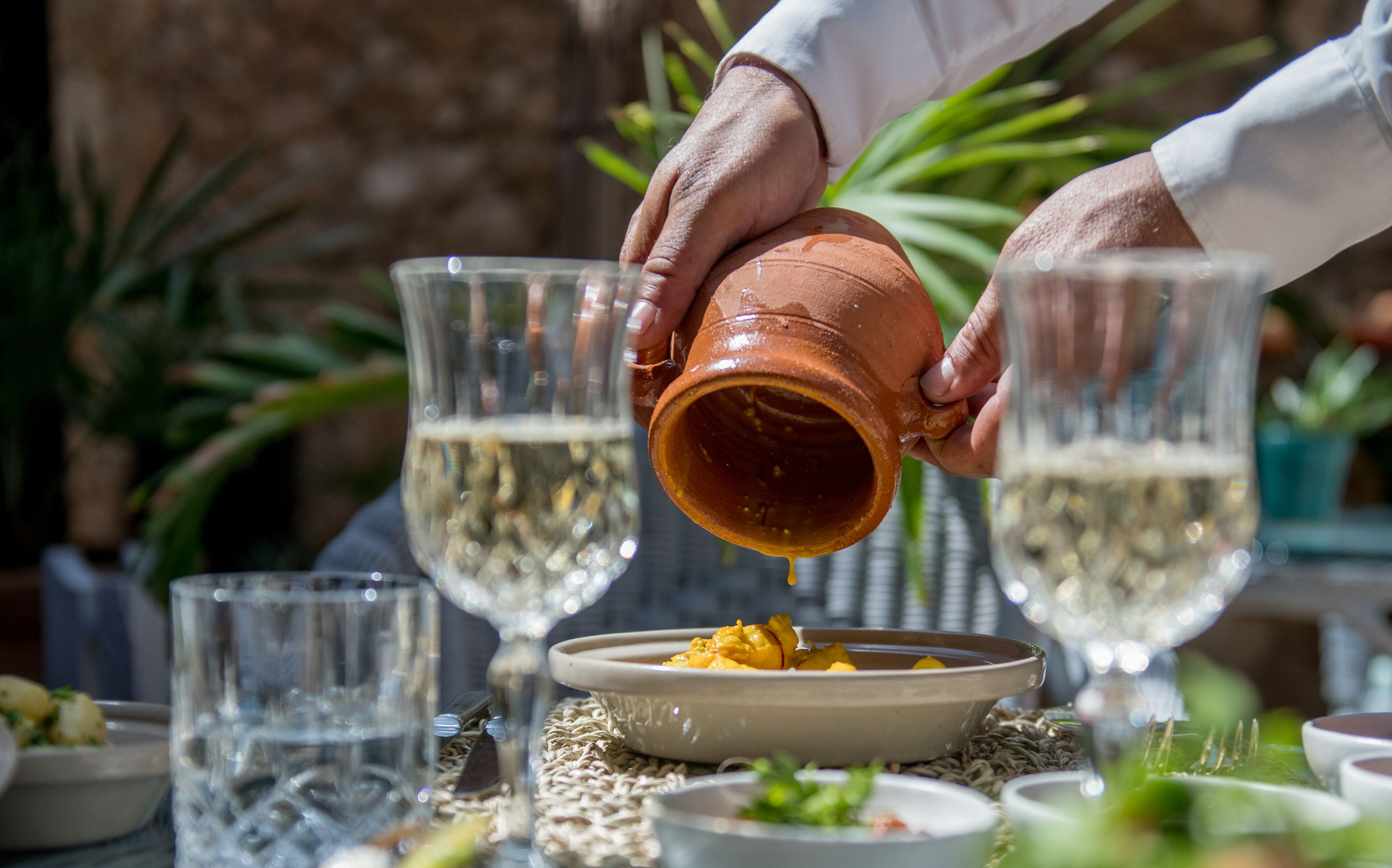 A person pouring a liquid from a clay pot into a bowl, with glasses and other dishes on the table.