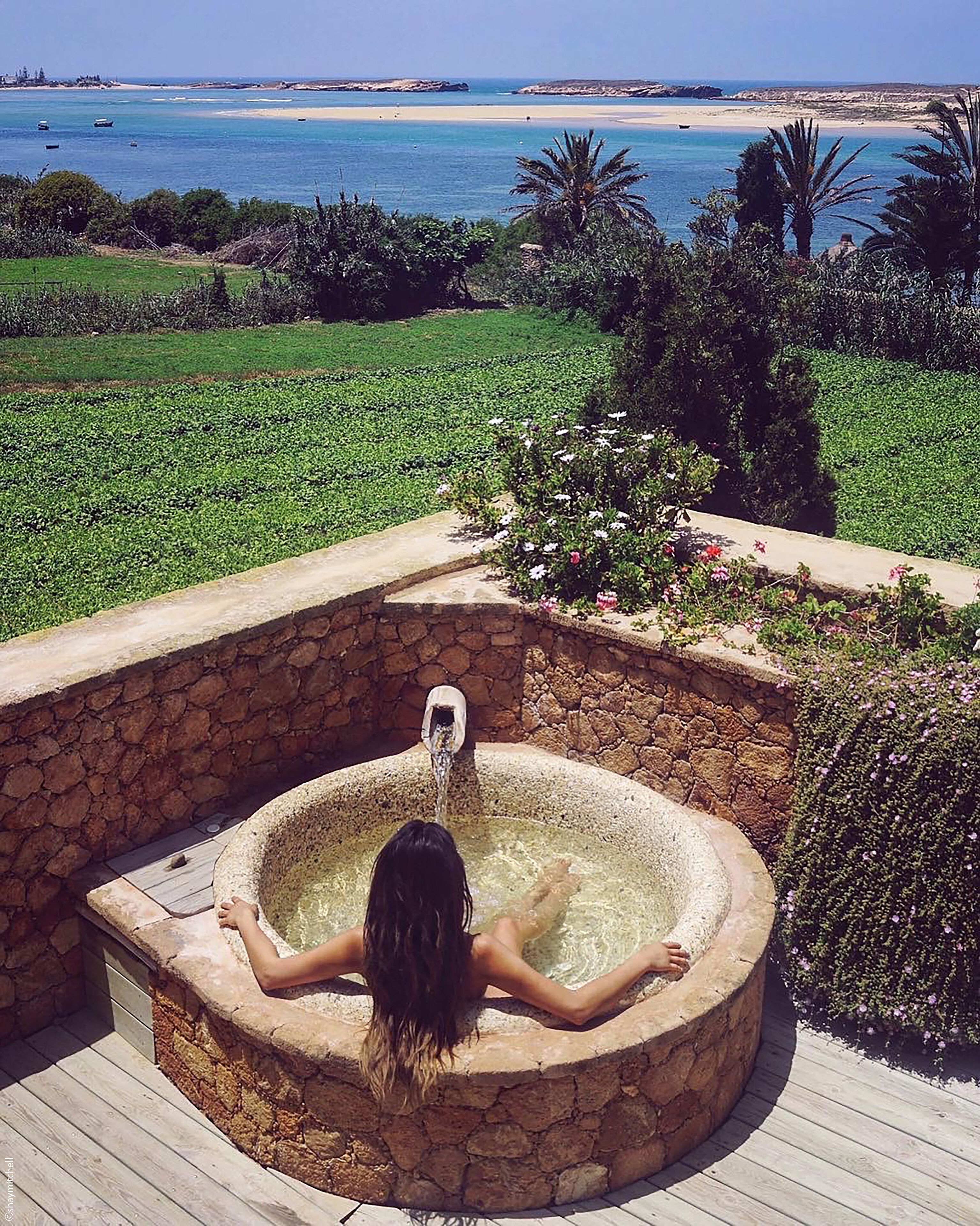 A person relaxing in a stone hot tub with a view of a beach and ocean.