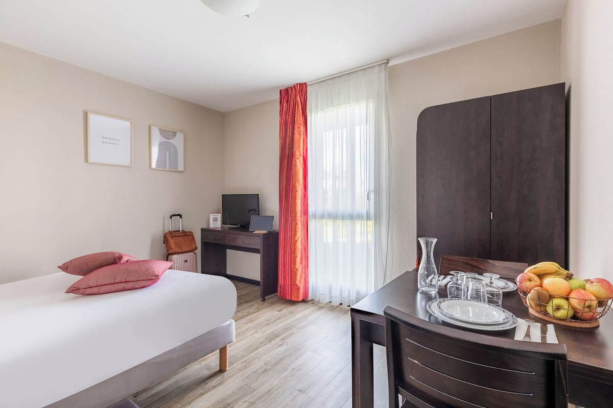 A hotel room with a bed, a desk with a laptop, a TV, a dining table with a fruit bowl, and a wardrobe.