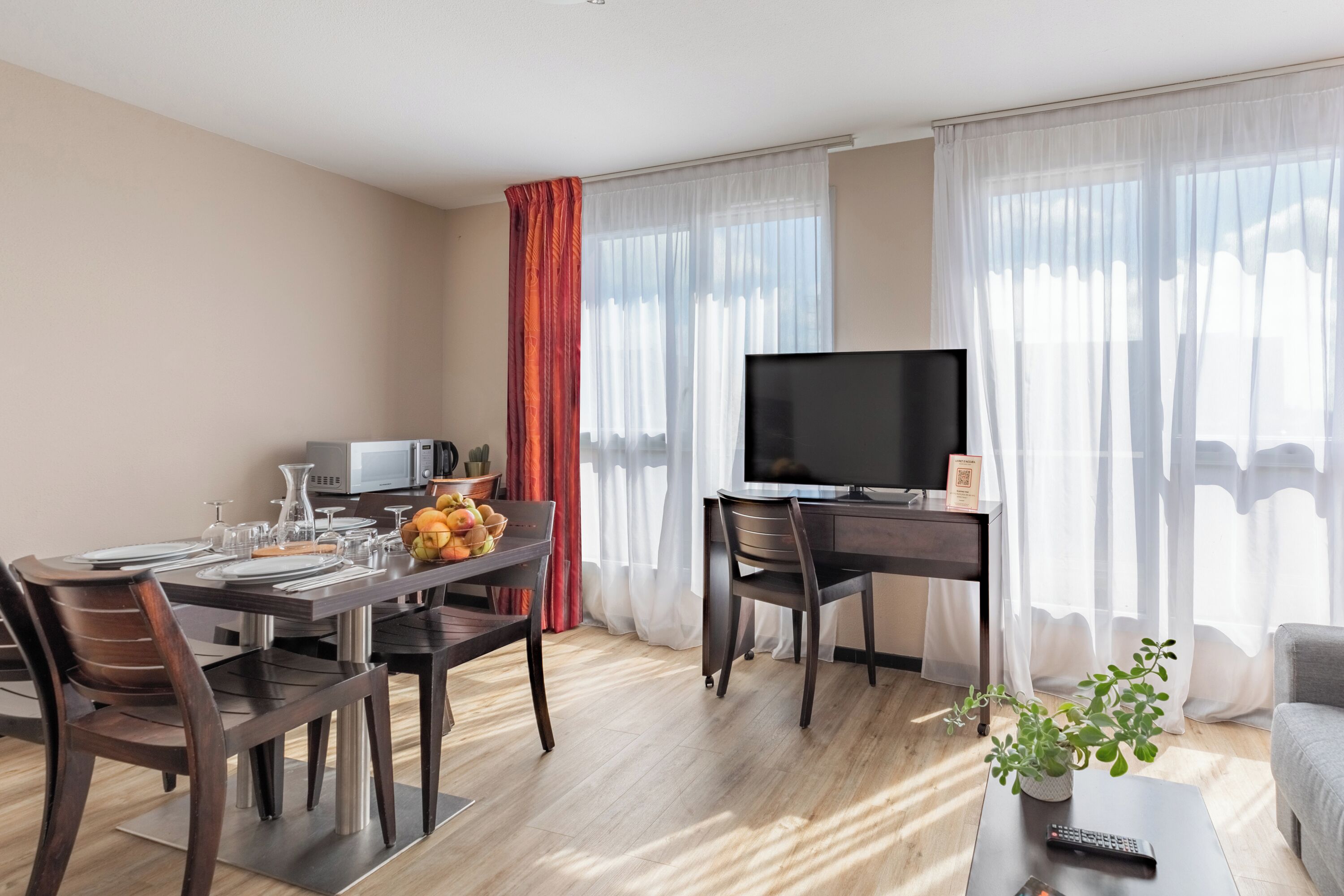 A modern hotel room with a dining area, a flat-screen TV, a sofa, and a potted plant.