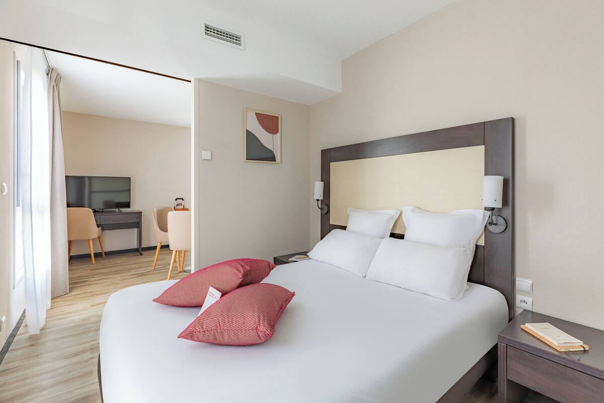 A hotel room with a large bed, two red pillows, a bedside table with a lamp, a desk with a chair, and a television.